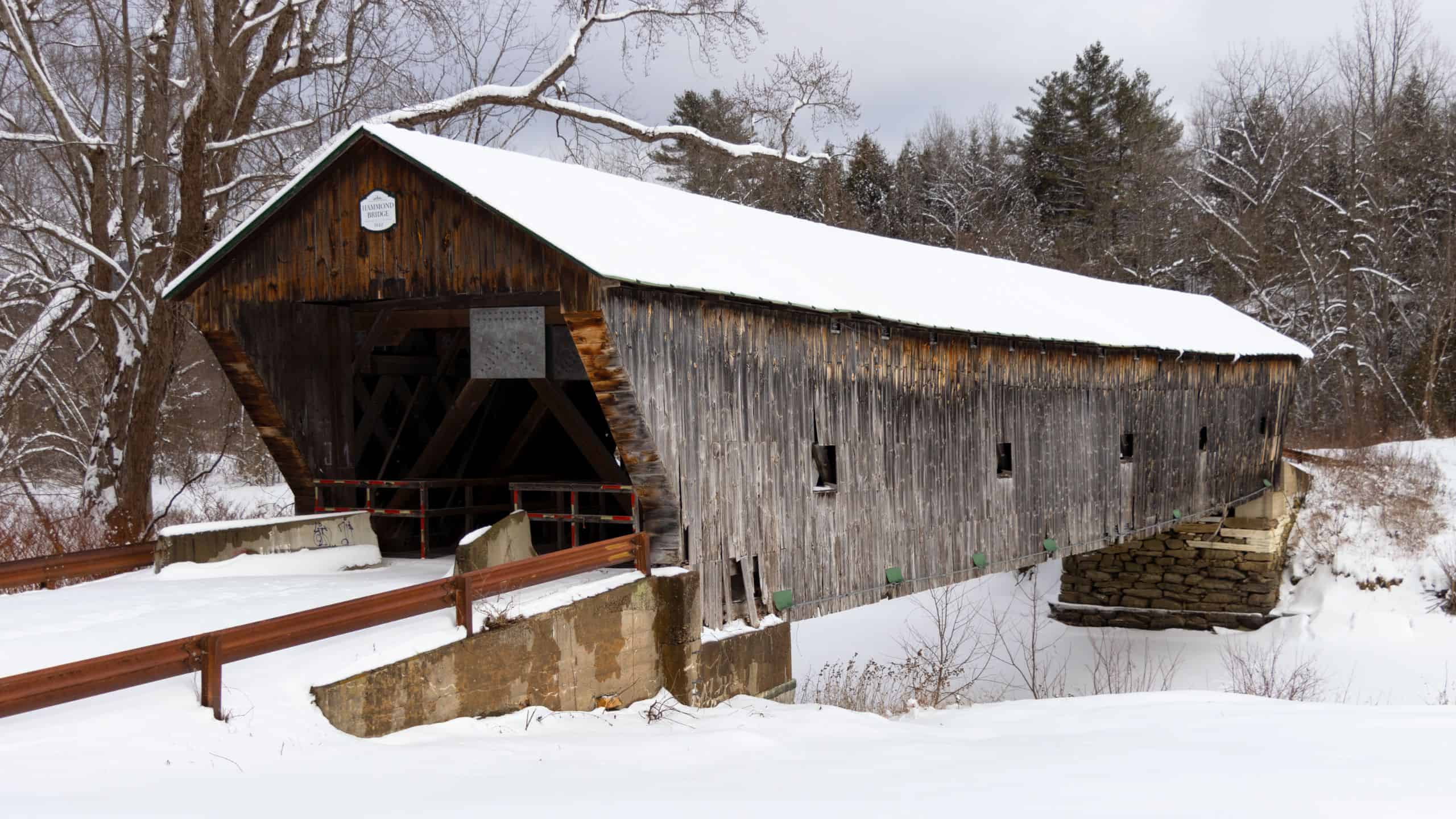 A wooden covered bridge spans a snow-covered river with bare trees and an overcast sky in the background.