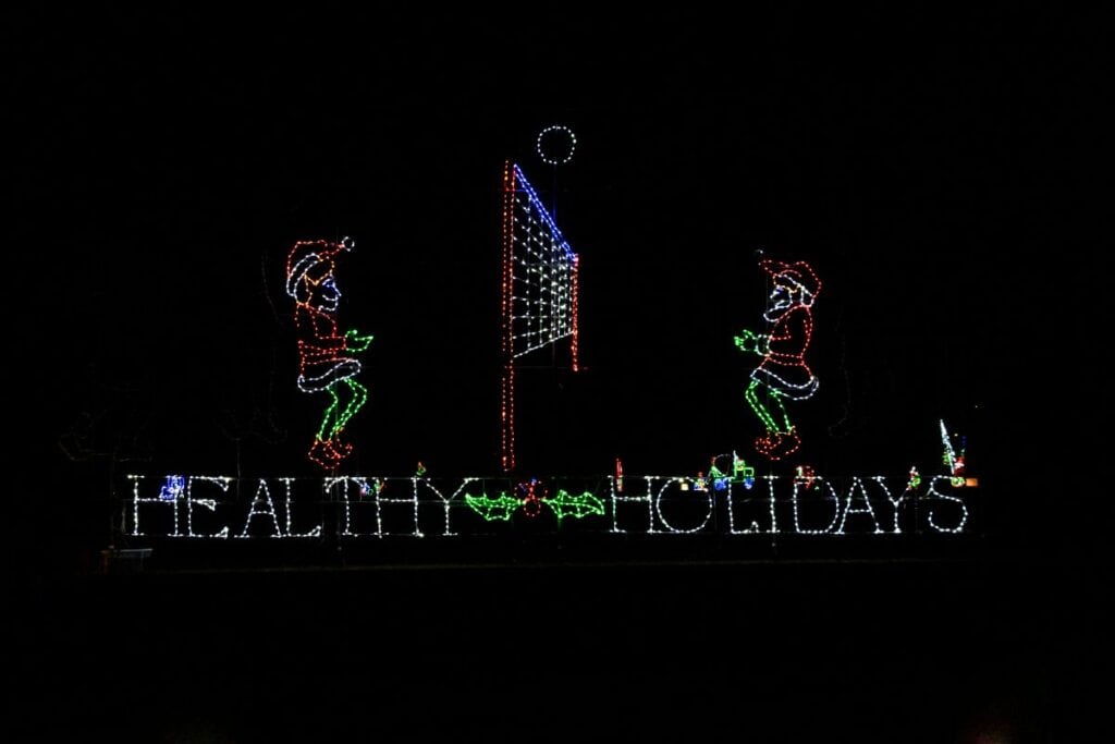 Holiday light display showing two elves playing volleyball above the words "Healthy Holidays" with holly decorations in between.