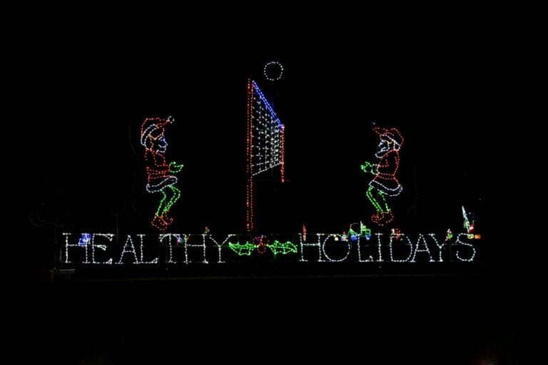 Holiday light display showing two elves playing volleyball above the words "Healthy Holidays" with holly decorations in between.