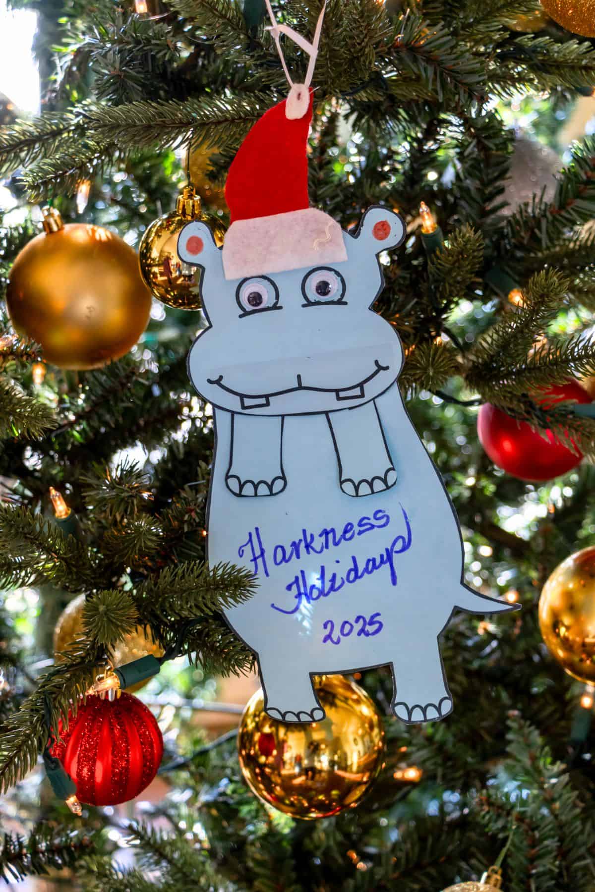 A blue paper hippopotamus ornament wearing a red Santa hat hangs on a Christmas tree with gold and red baubles. The ornament reads, "Harkness Holiday 2023.