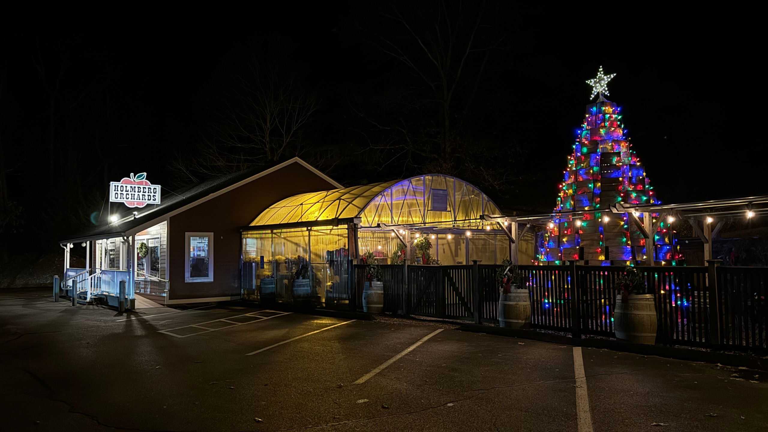 A building with a lit "Holmdel Orchards" sign is next to a large Christmas tree decorated with colorful lights at night. The area appears empty.