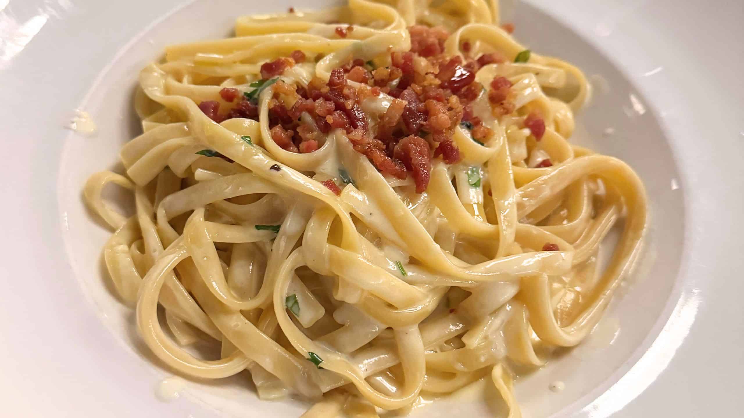 A plate of fettuccine pasta topped with creamy sauce, chopped herbs, and crispy bacon bits.