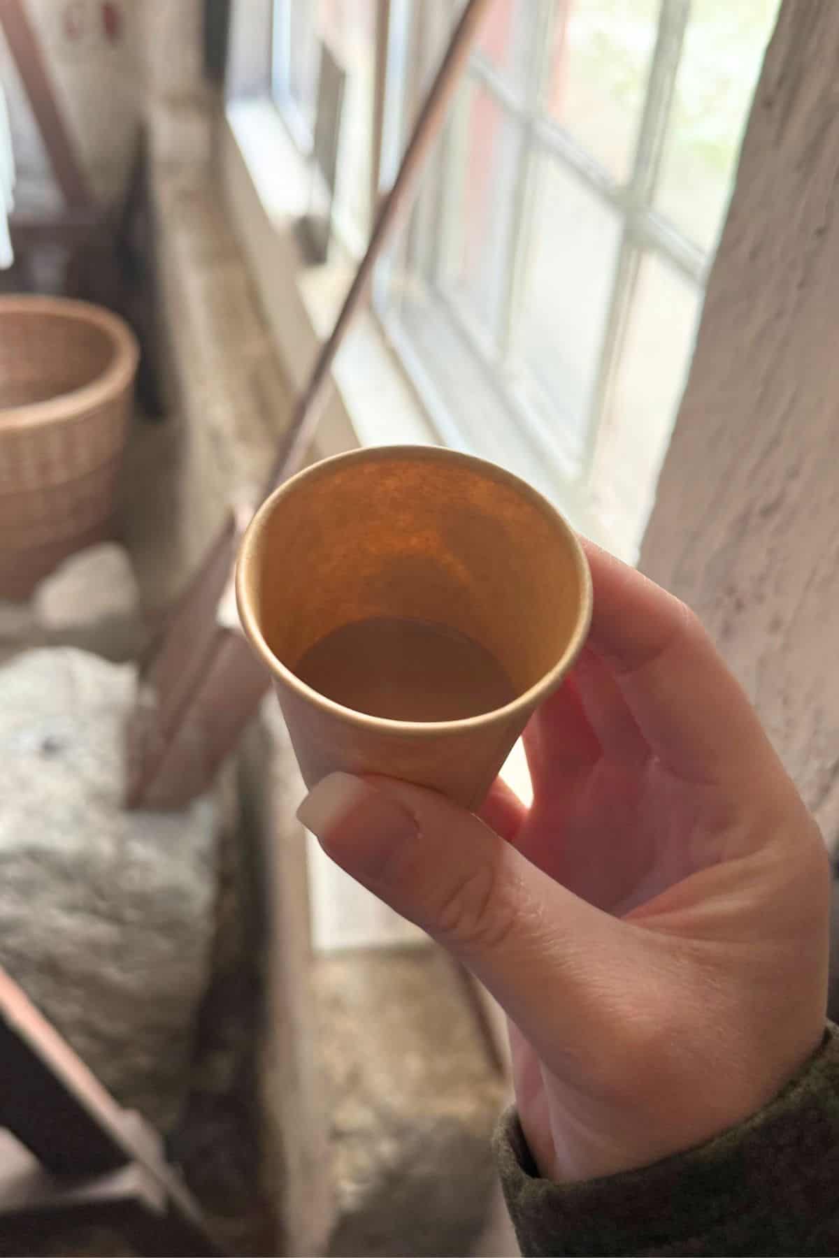 A hand holds a small paper cup with a brown liquid inside, near a window with stone and wicker basket visible in the background.