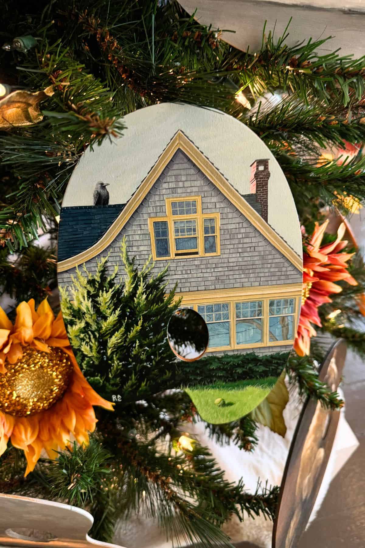 A painted artist's palette depicting a gray house with yellow trim hangs as an ornament on a decorated Christmas tree with lights and artificial sunflowers.