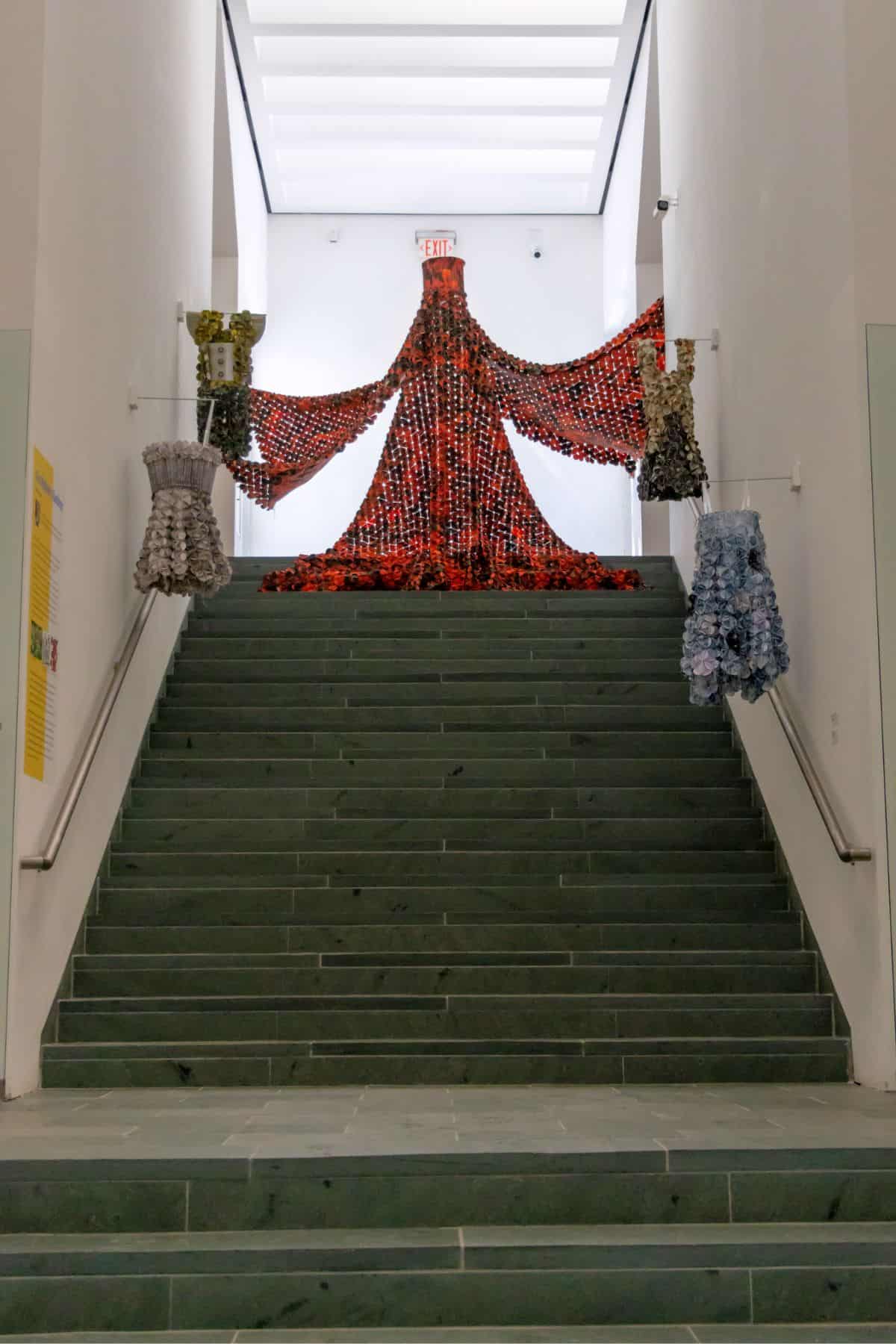 A large, red robe-like sculpture is displayed at the top of a staircase, flanked by two smaller, textured dress sculptures on either side of the stairway.