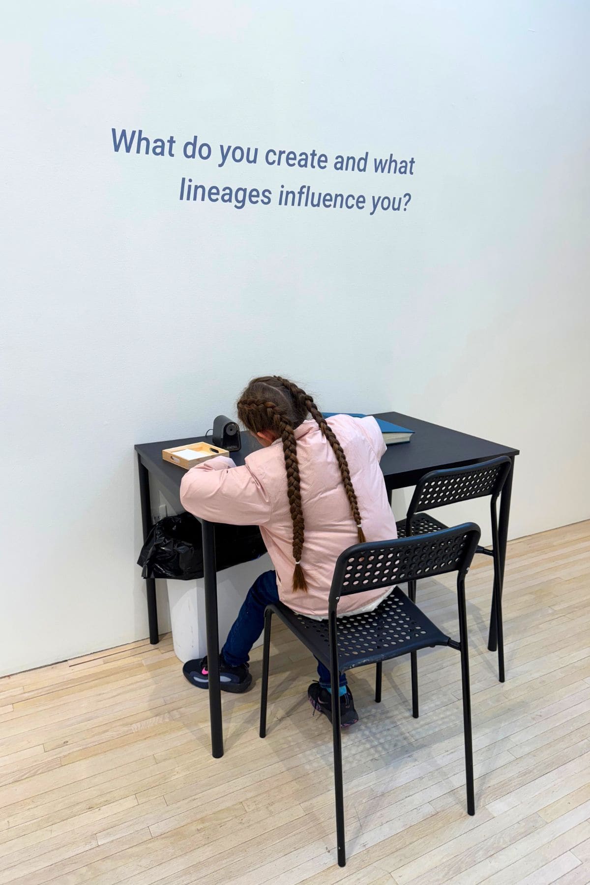 A girl with braided hair sits at a black table writing, beneath a wall sign that reads, "What do you create and what lineages influence you?.