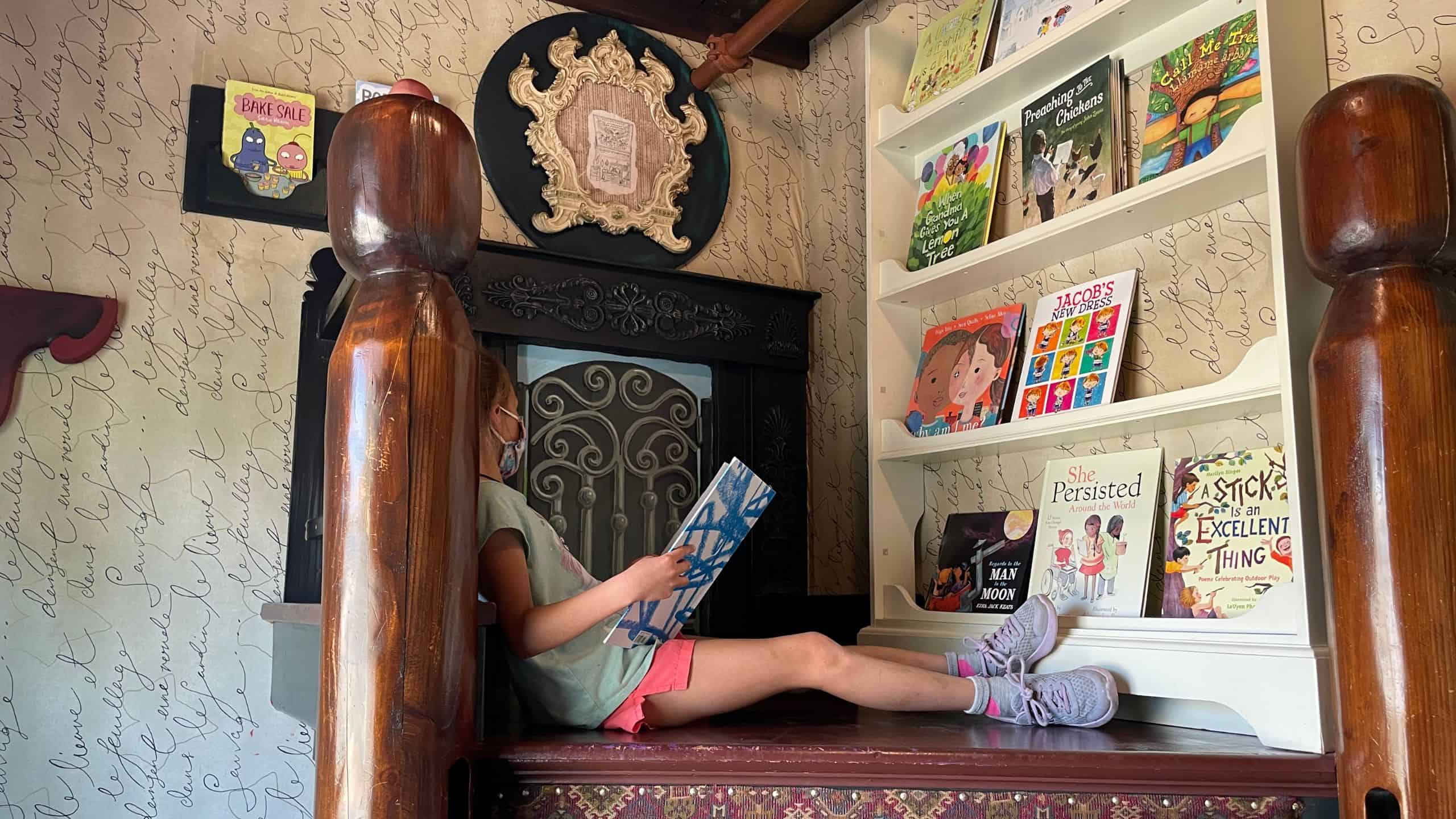 A child sits on a wooden ledge reading a book in a cozy room with shelves displaying various children's books.