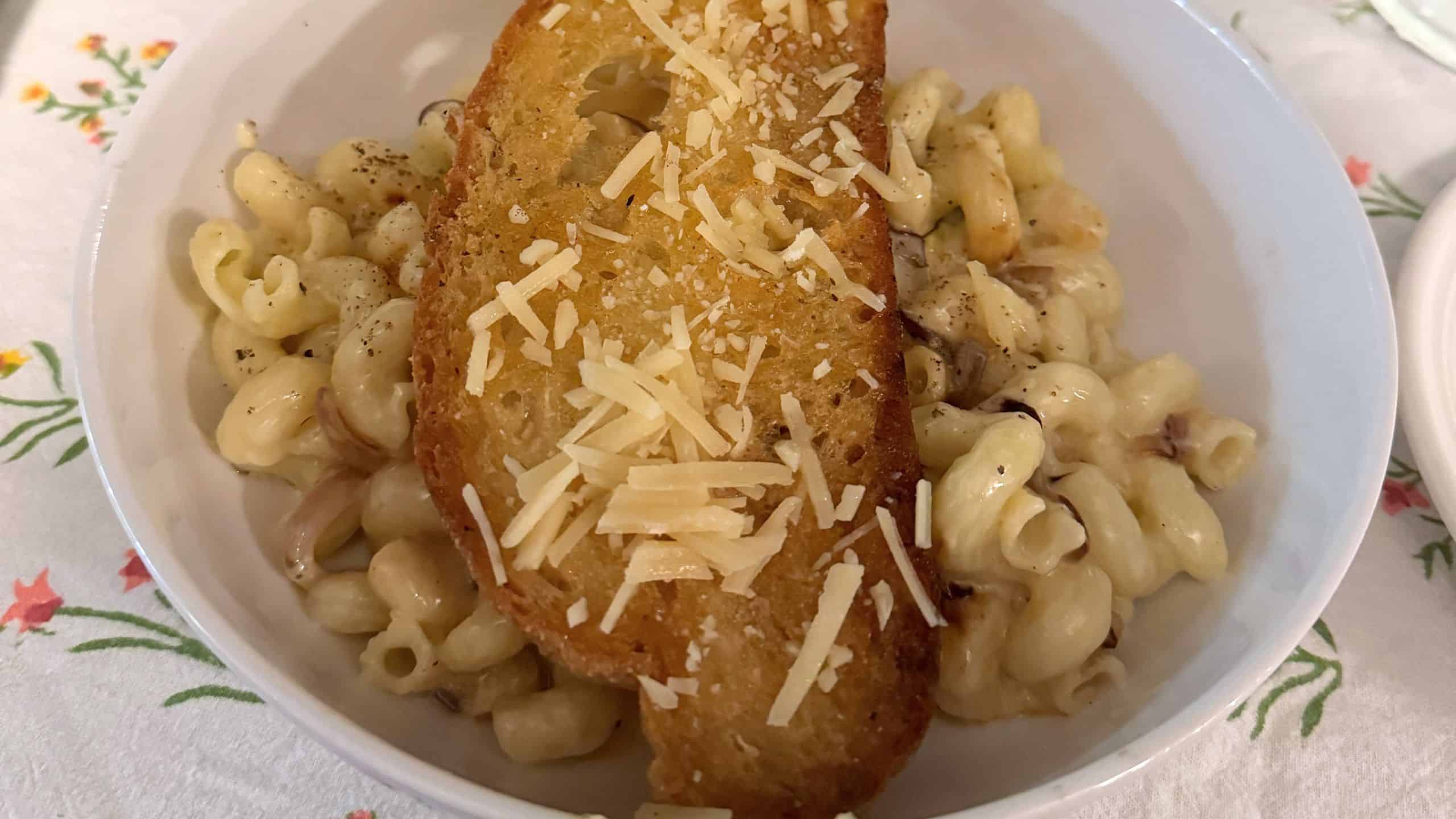A bowl of macaroni and cheese topped with a slice of toasted bread and grated cheese, placed on a floral tablecloth.