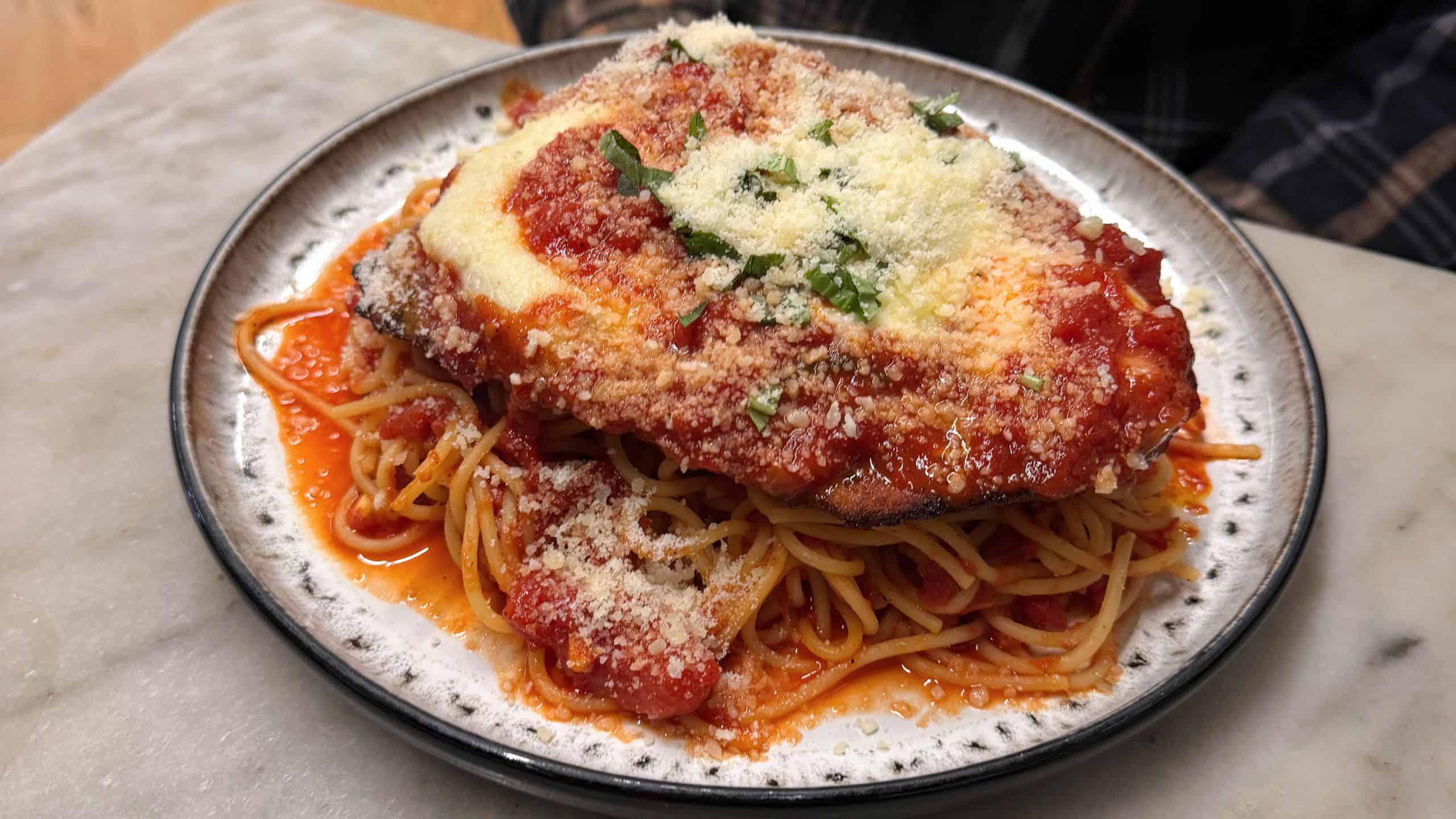 A plate of spaghetti topped with breaded chicken, marinara sauce, melted cheese, grated parmesan, and chopped herbs.