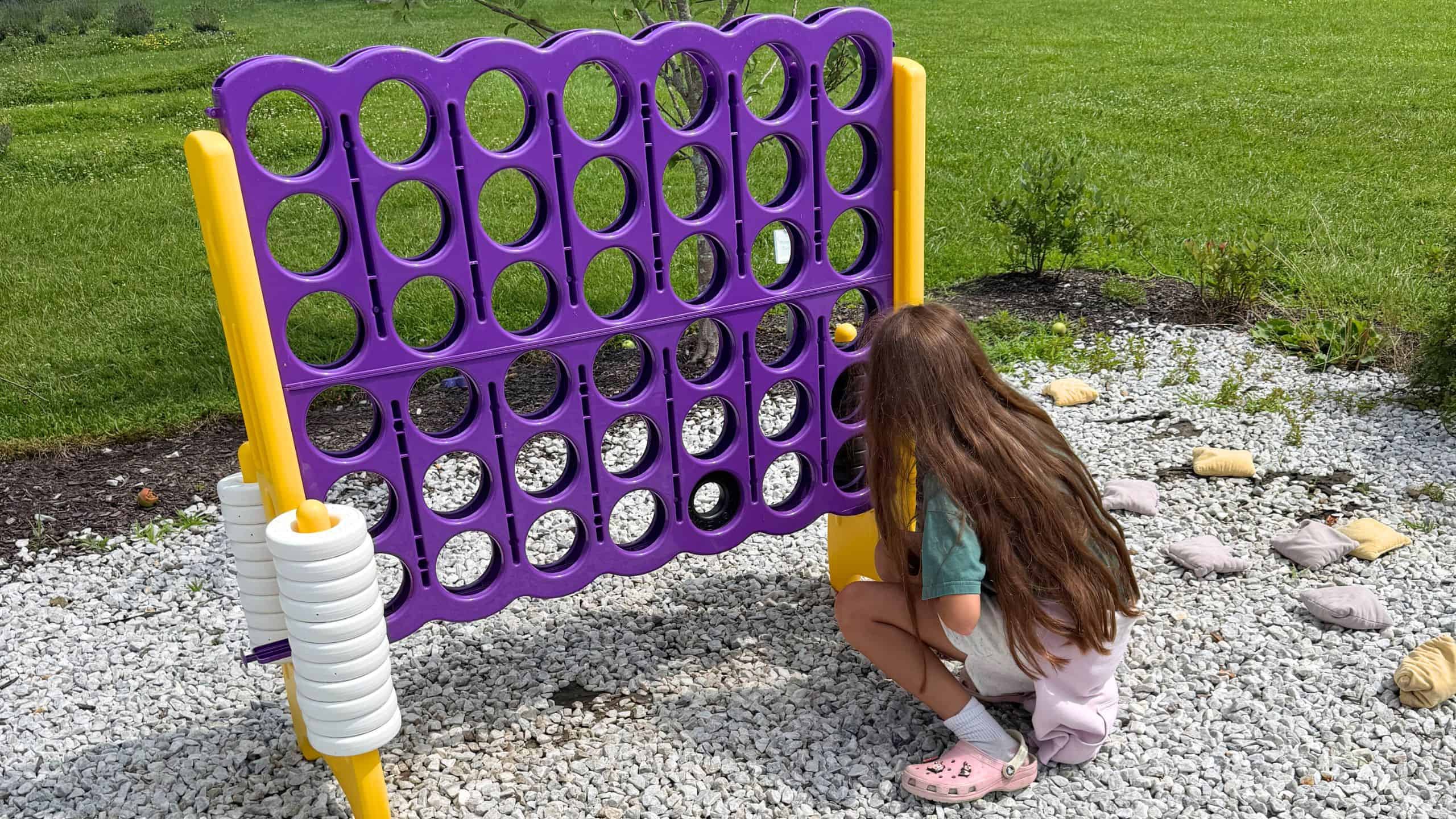 A girl with long brown hair plays a large outdoor Connect Four game on a gravel area with green grass in the background.