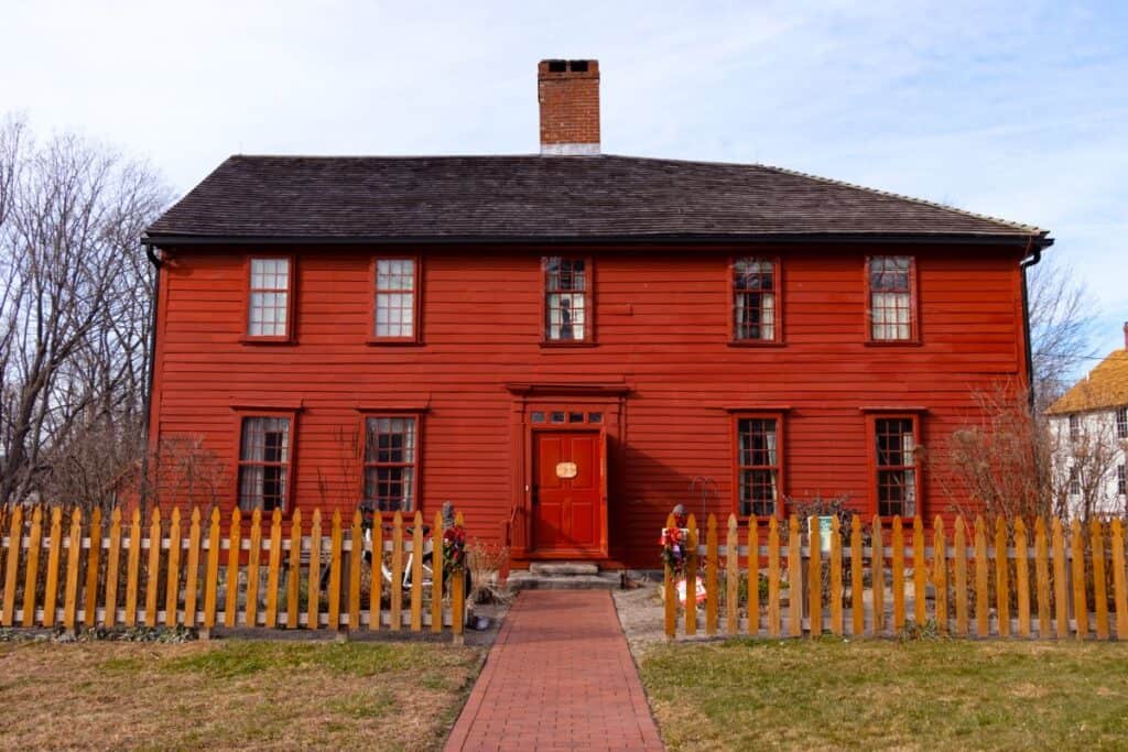 A two-story red wooden house with a central brick chimney, a picket fence, and a brick path leading to the front door, photographed on a clear day.