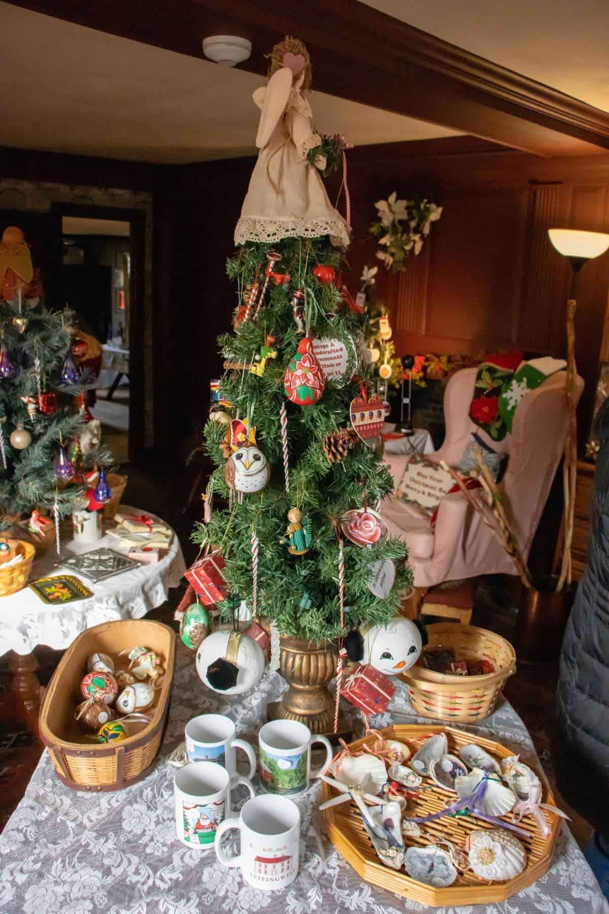 A small decorated Christmas tree with ornaments is displayed on a table with mugs, baskets of seashells, and holiday-themed items in a warmly lit room.