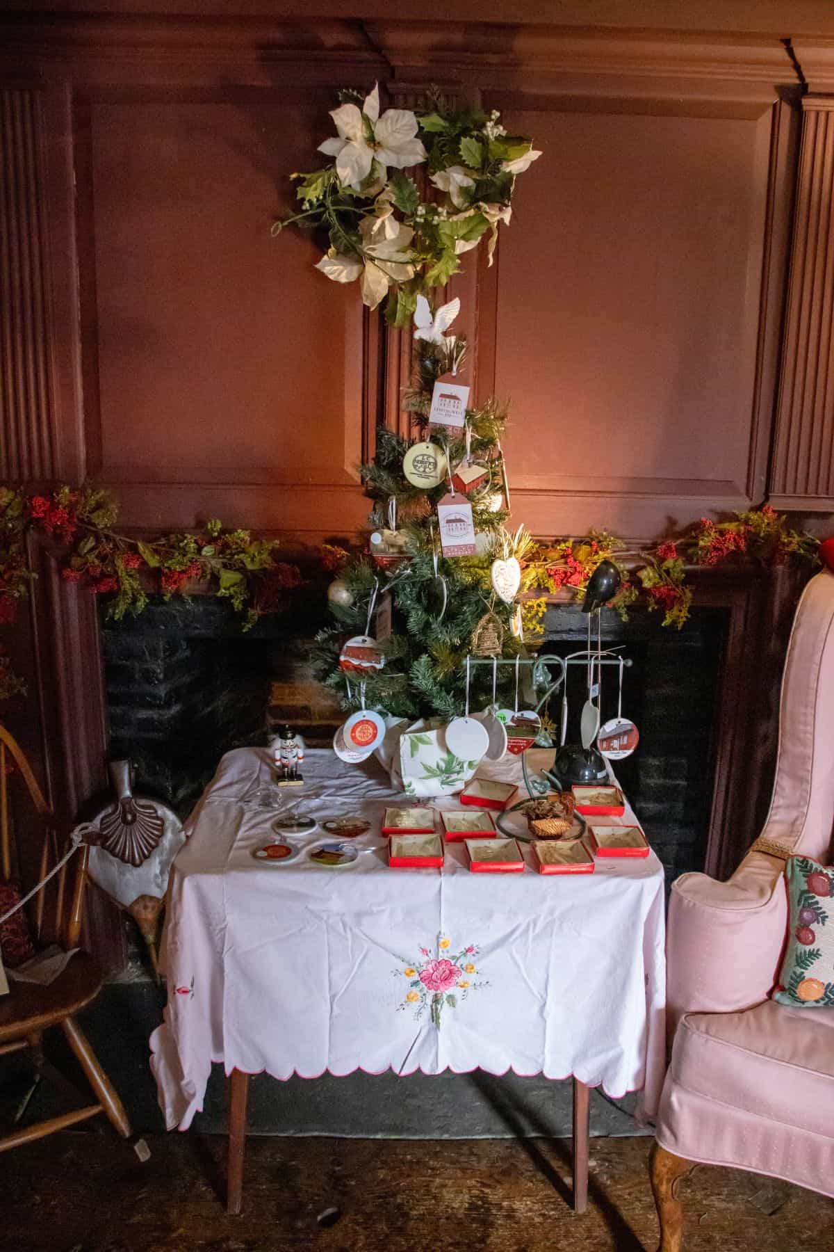 A decorated small Christmas tree stands on a fireplace mantel behind a table set with books, cups, snacks, and a white embroidered tablecloth in a cozy room.