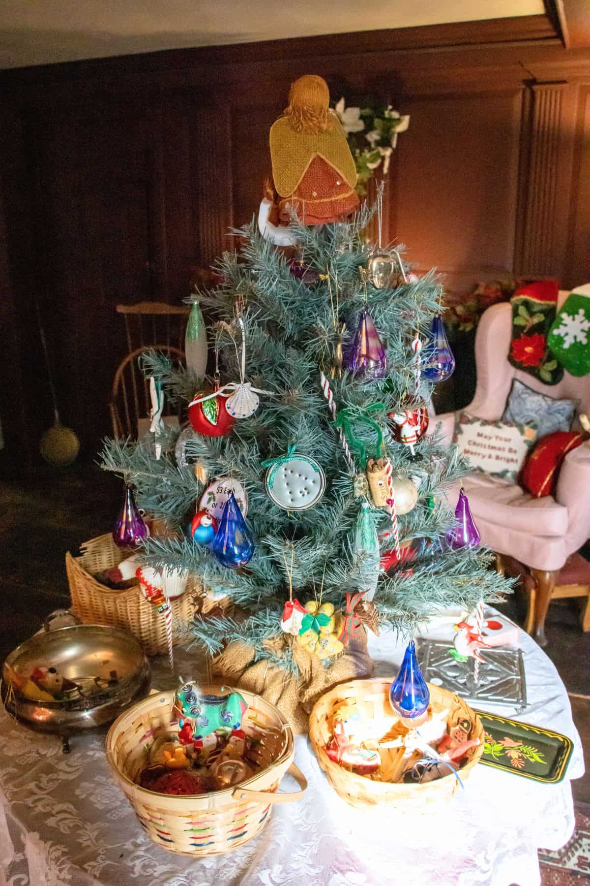 A small decorated Christmas tree with various ornaments sits on a table, surrounded by baskets filled with decorations in a cozy room.