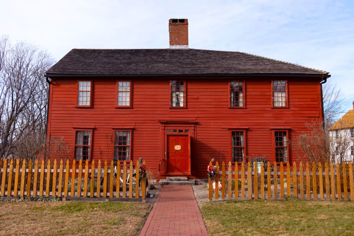 A two-story red wooden house with a central brick chimney, a picket fence, and a brick path leading to the front door, photographed on a clear day.