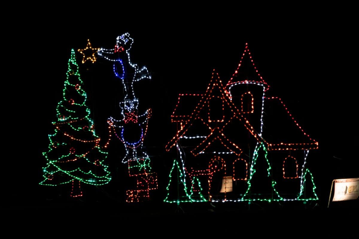 Christmas light display showing three snowmen stacking to place a star on a tree, next to a house outlined in lights, all against a black background.