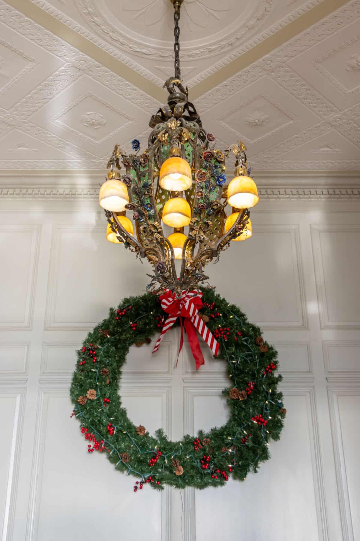 A chandelier hangs from a decorative ceiling above a large green wreath with red berries and a red striped ribbon, attached to a paneled wall.