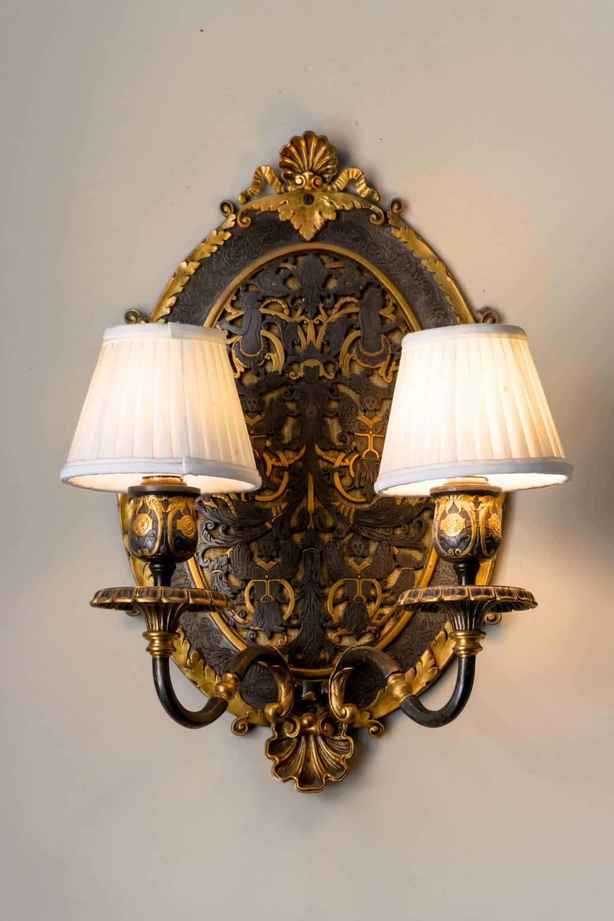 Ornate antique wall sconce with two lit lampshades, gold and black detailing, mounted on a white wall.
