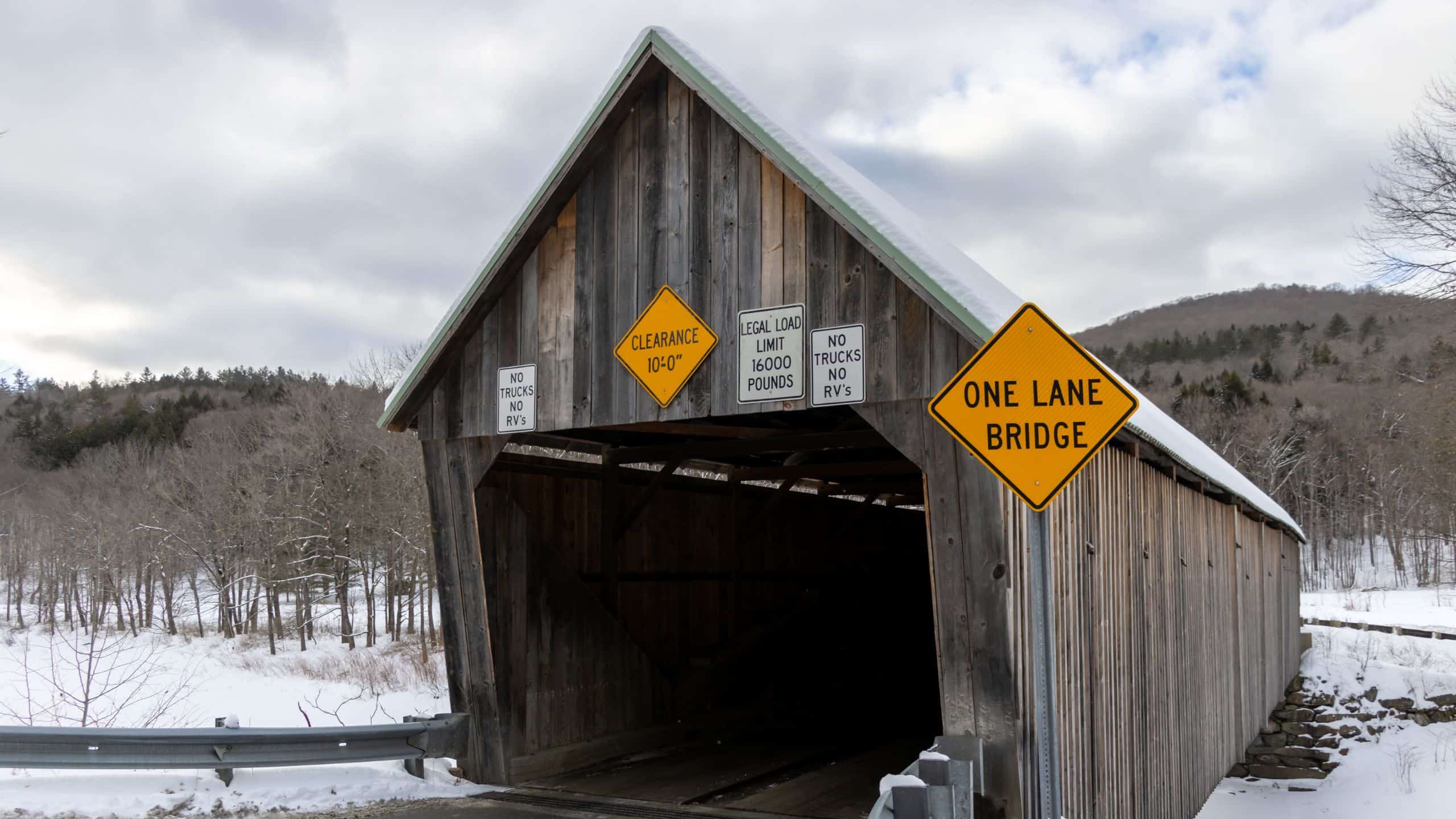 A classic New England covered bridge spans a snowy landscape, featuring signs for clearance height, weight limit, no trucks or RVs, and warning travelers it is a one-lane crossing.