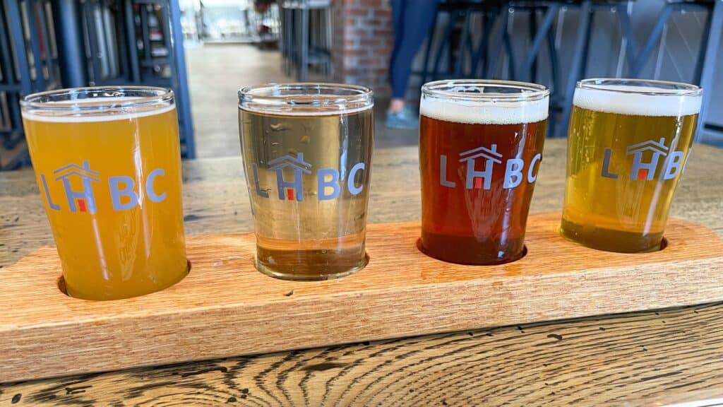 A wooden flight tray holds four small glasses of different colored beers, each labeled with the LHBC logo, on a wooden table in a brewery.