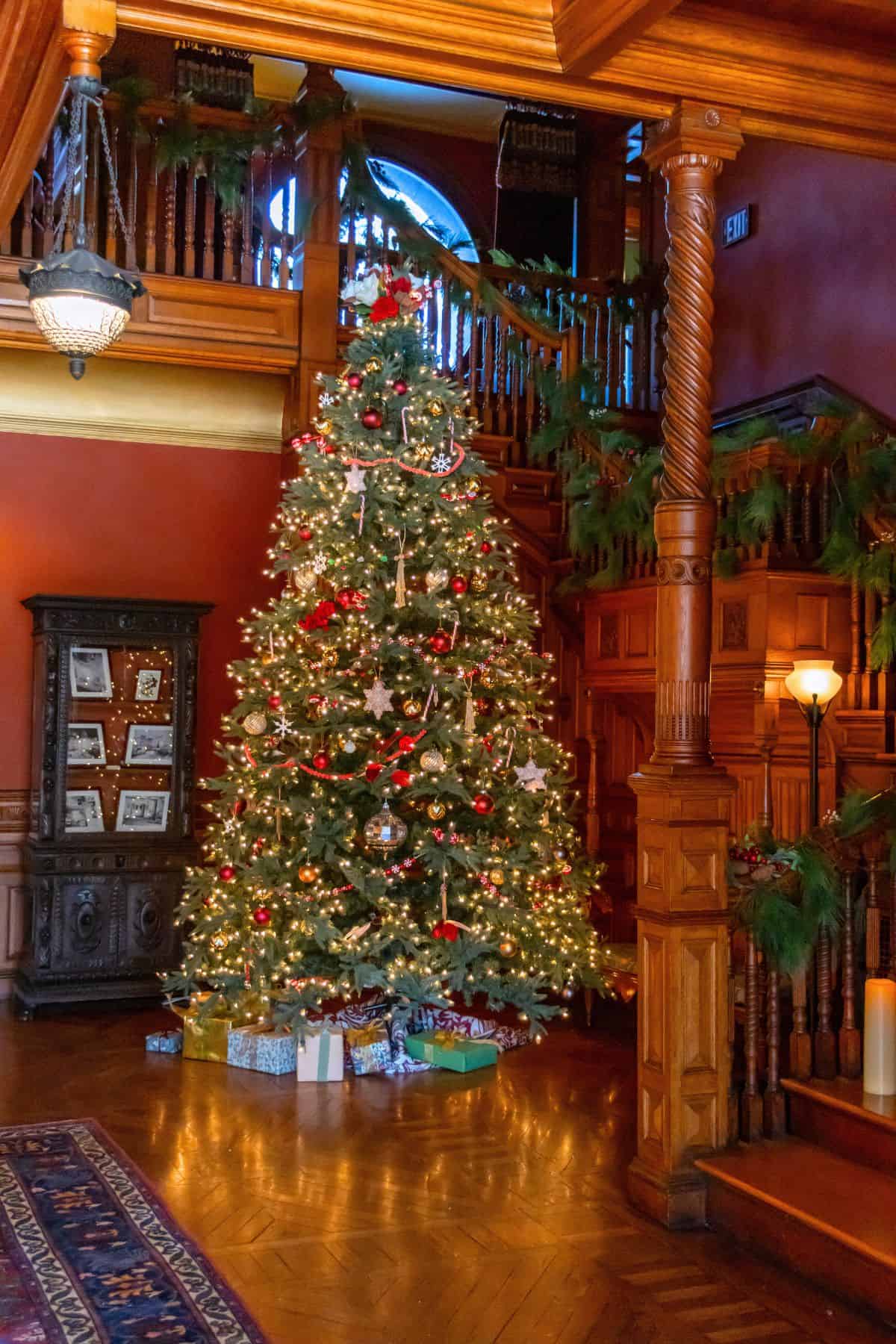 A decorated Christmas tree with lights, ornaments, and wrapped presents stands in the corner of a wooden-paneled room with a staircase and festive greenery.