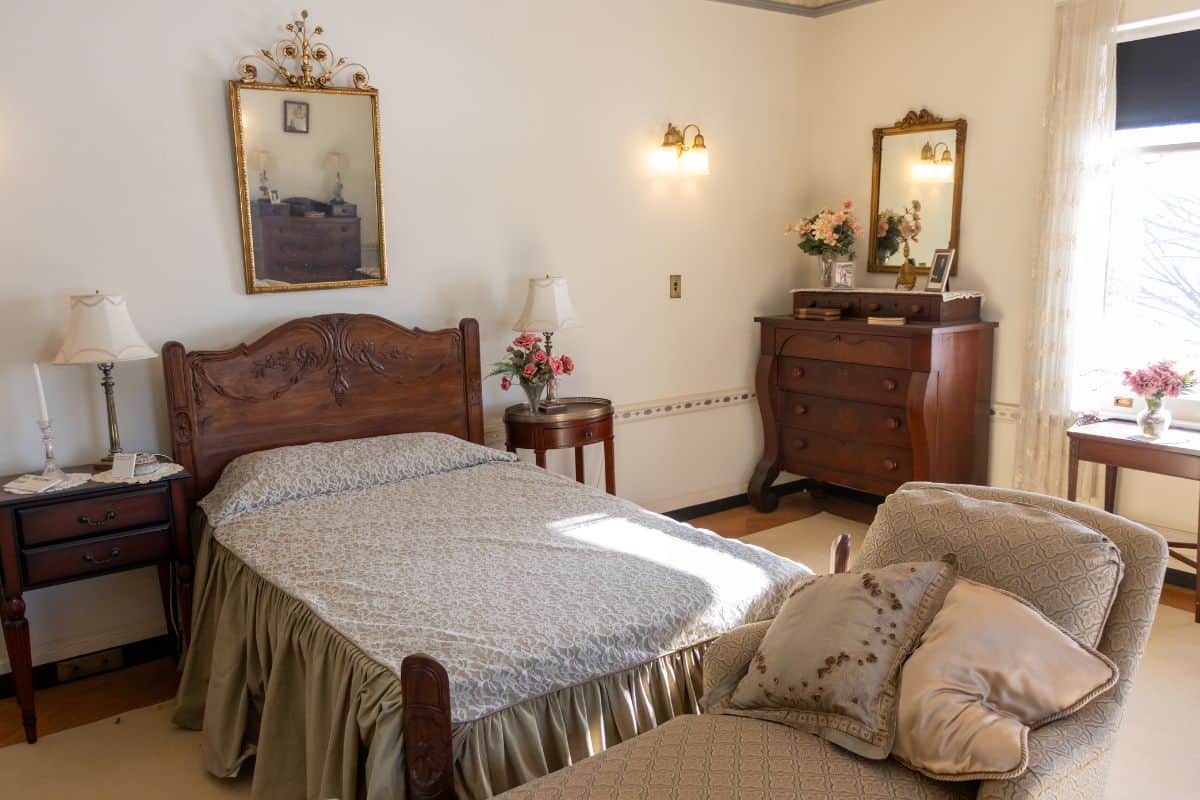 A neatly made bed with a carved wooden headboard, a side table, a chaise lounge with pillows, and a dresser with a mirror in a well-lit, traditional bedroom.