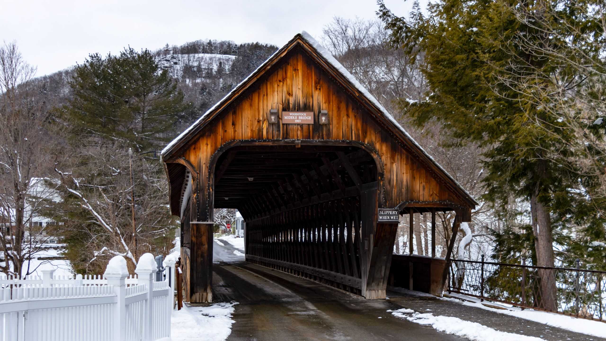 A wooden covered bridge crosses a snowy road in a winter landscape, surrounded by trees and hills in the background.