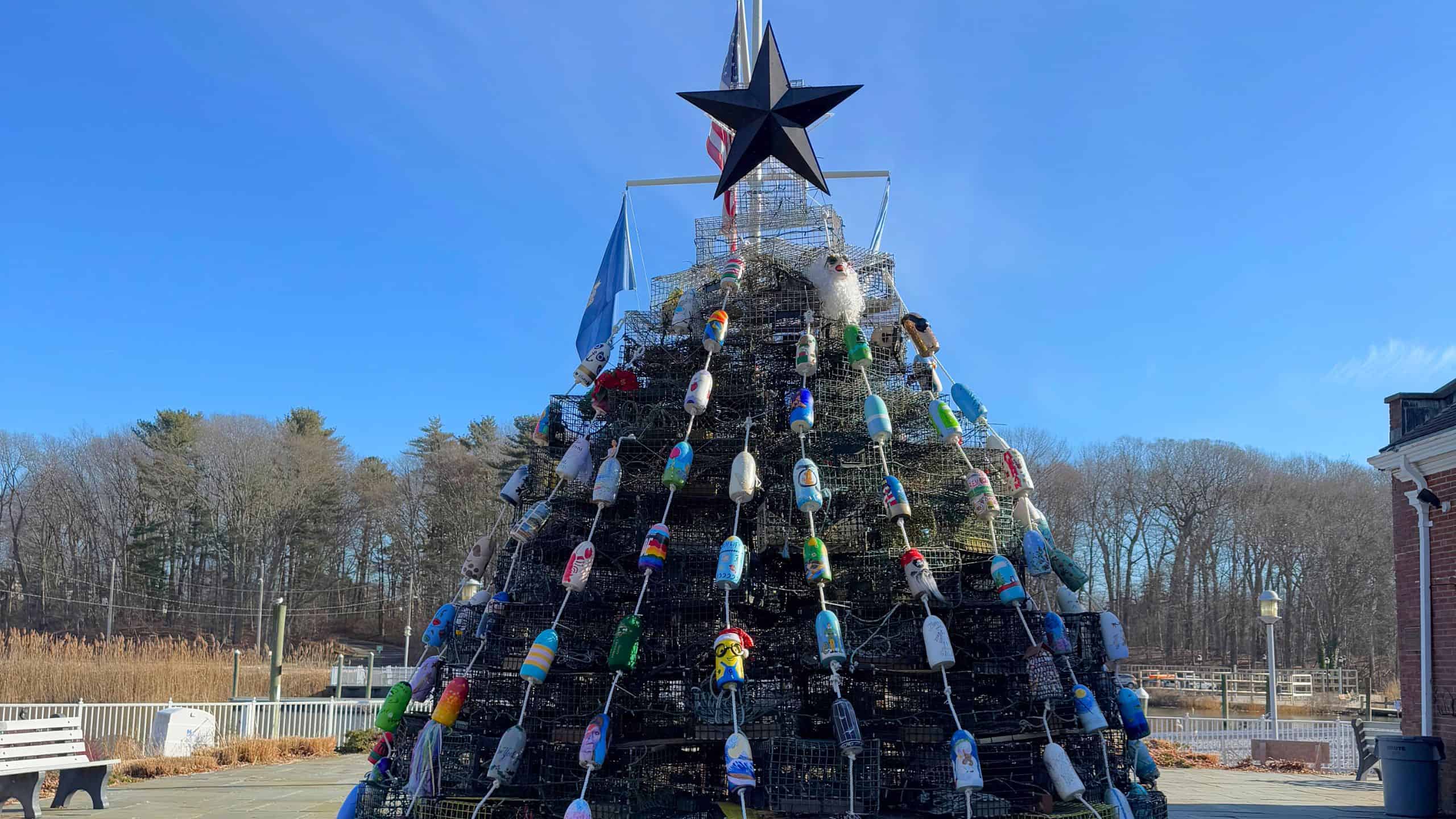 A large outdoor tree-shaped structure made of stacked metal cages and decorated with colorful plastic buoys, topped with a black star, stands in a paved area near trees and buildings.