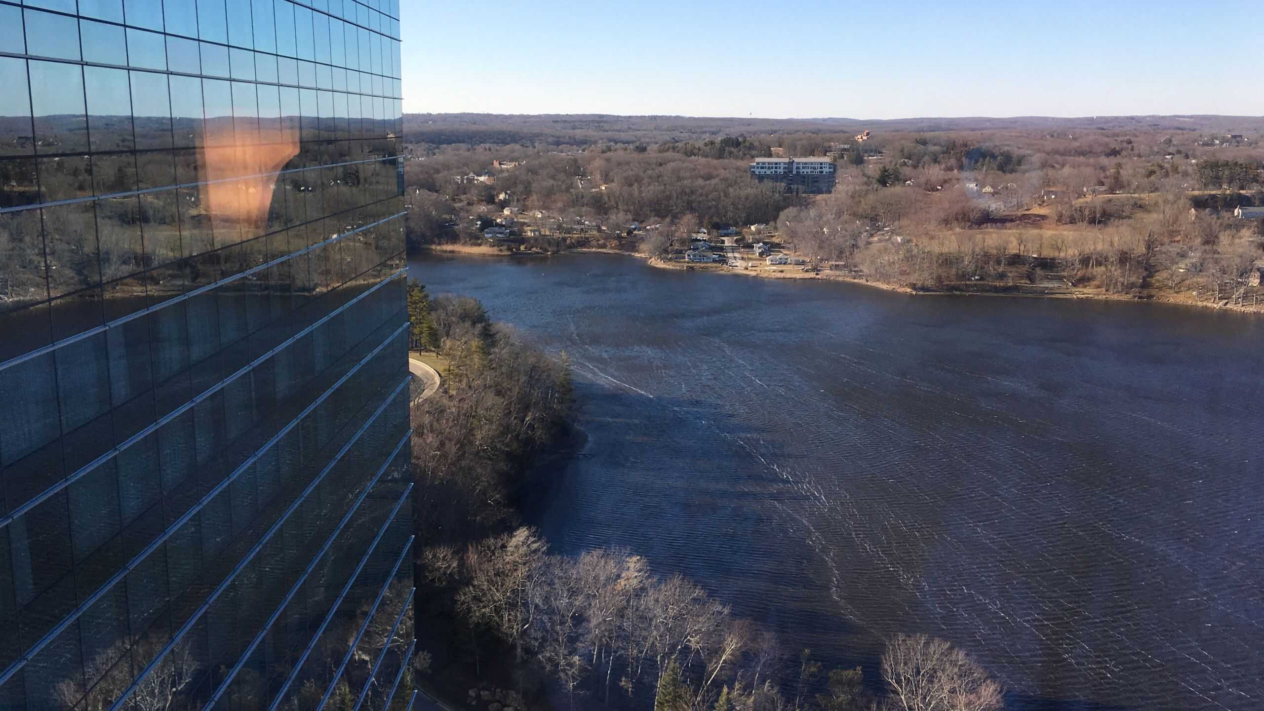 High-rise glass building reflects sunlight next to a lake, with leafless trees and distant houses under a clear blue sky.