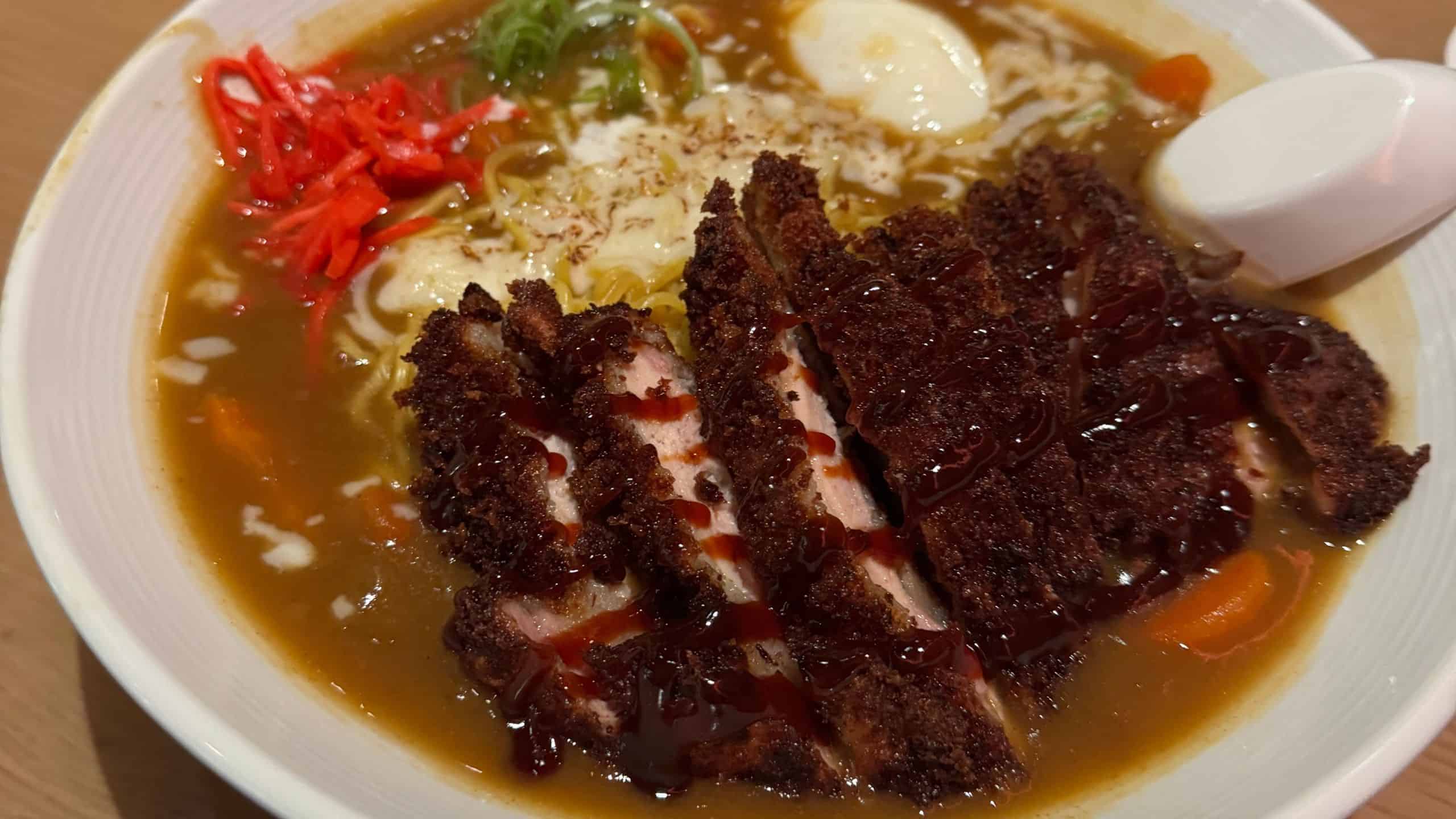 A bowl of ramen topped with sliced breaded pork cutlet, curry sauce, pickled ginger, green onions, and a spoon.