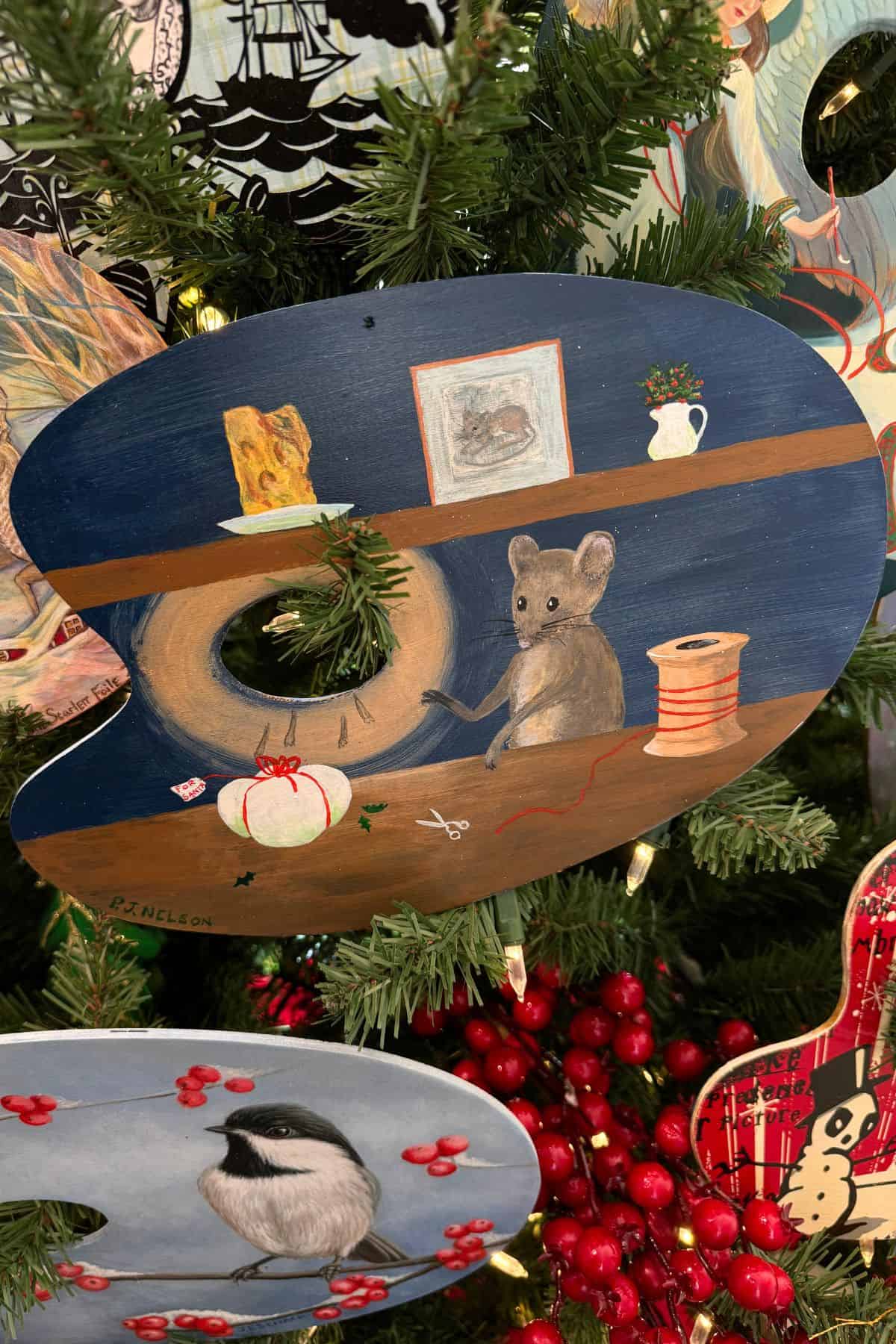 A painted wooden artist's palette shows a mouse sitting beside a spool of thread, a piece of cheese, a framed picture, and a vase on a shelf, hanging on a decorated Christmas tree.