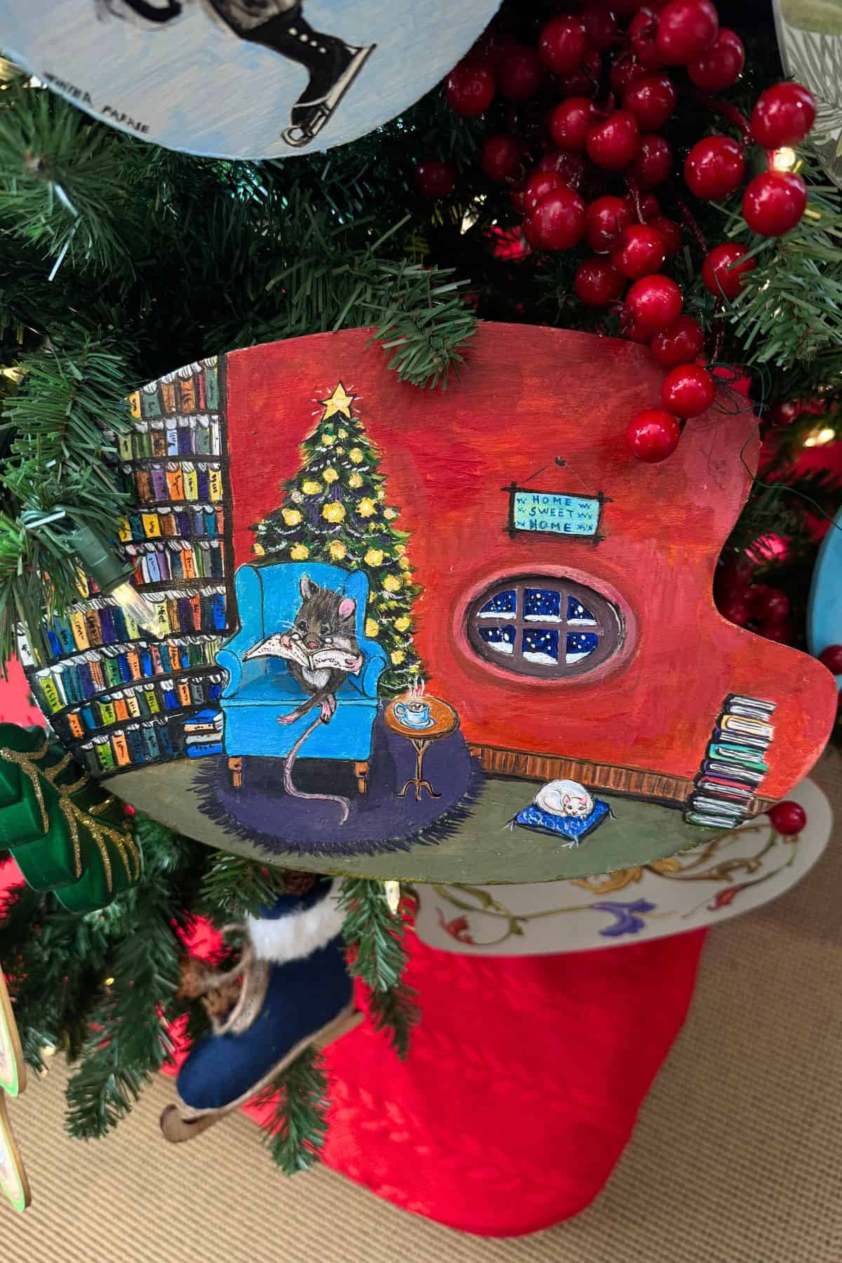 A painted palette shows a rat reading in an armchair beside a Christmas tree, with bookshelves, a window, and festive decorations in the background, hung on a decorated Christmas tree.