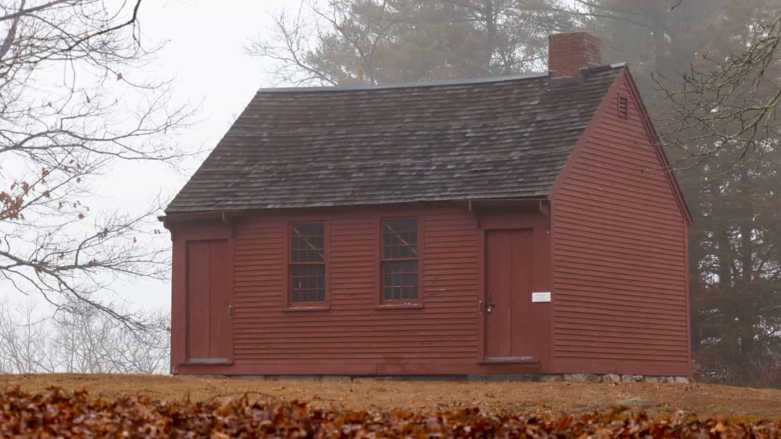 A small red wooden cabin, reminiscent of historic sites in Connecticut, features two central windows, wooden doors on each side, and a shingled roof, set on a foggy day with bare trees in the background.