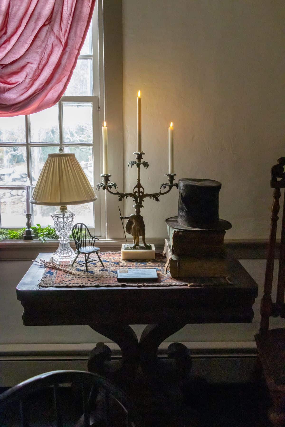 A wooden table by a window with a candelabra, a lamp, a miniature chair, stacked books, a top hat, and a small rug.