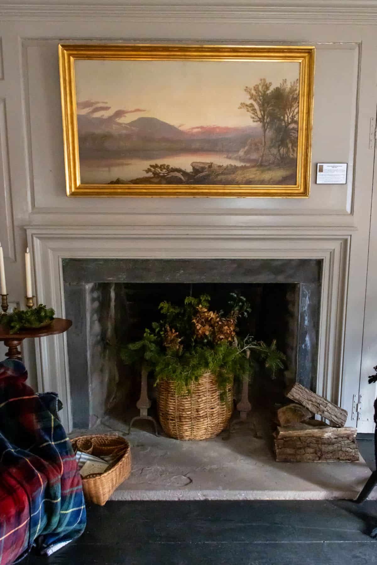 A fireplace with a basket of greenery inside, a stack of firewood beside it, a plaid blanket on a chair, and a landscape painting hanging above the mantel.