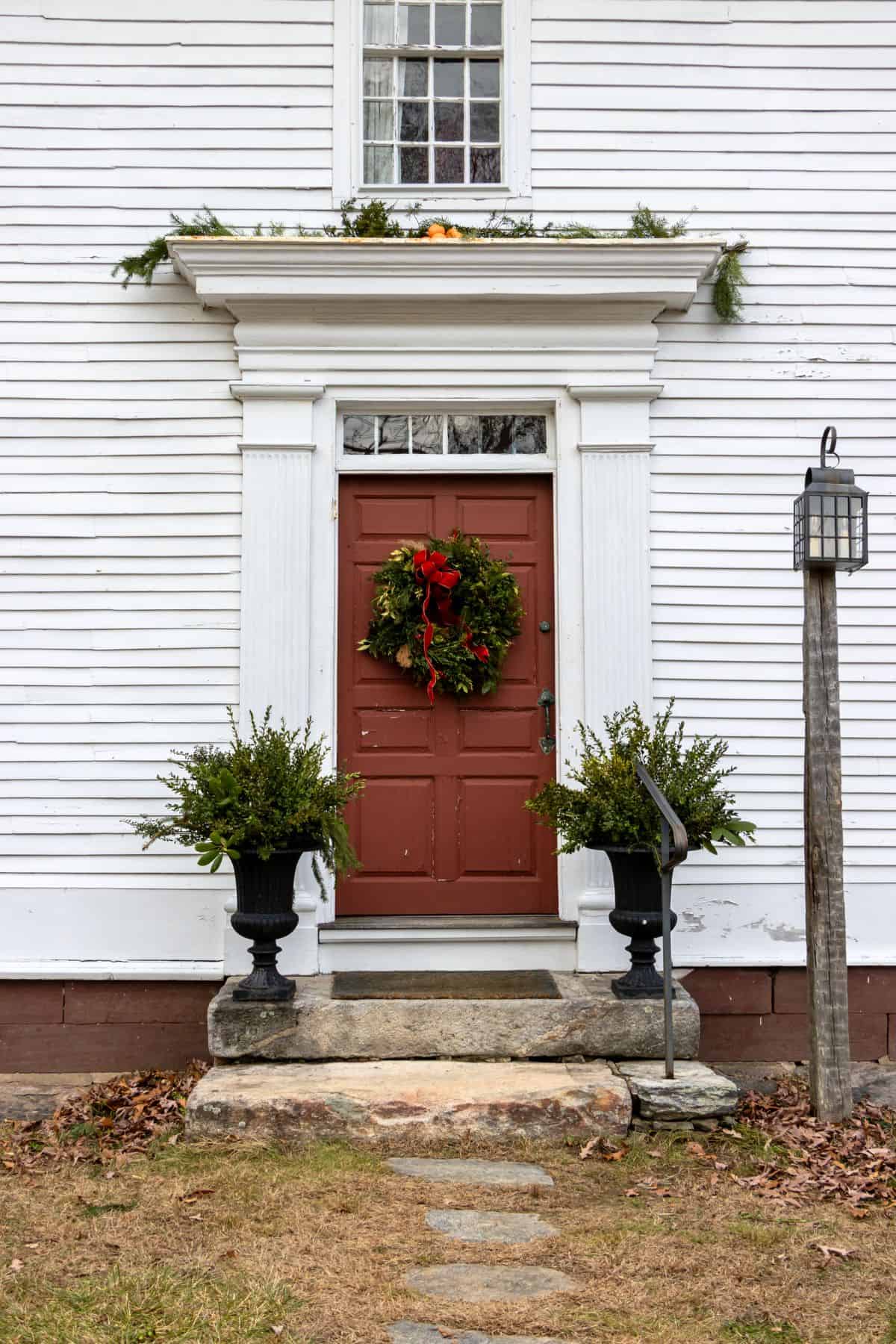 Red door with a holiday wreath flanked by two potted plants, set in a white wooden exterior with a stone step and a lantern on the right side.