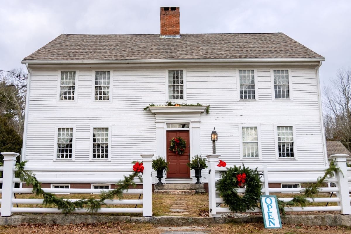 A large white colonial-style house with a red door, holiday wreaths, green garlands, and a sign that says "OPEN" by the front fence.