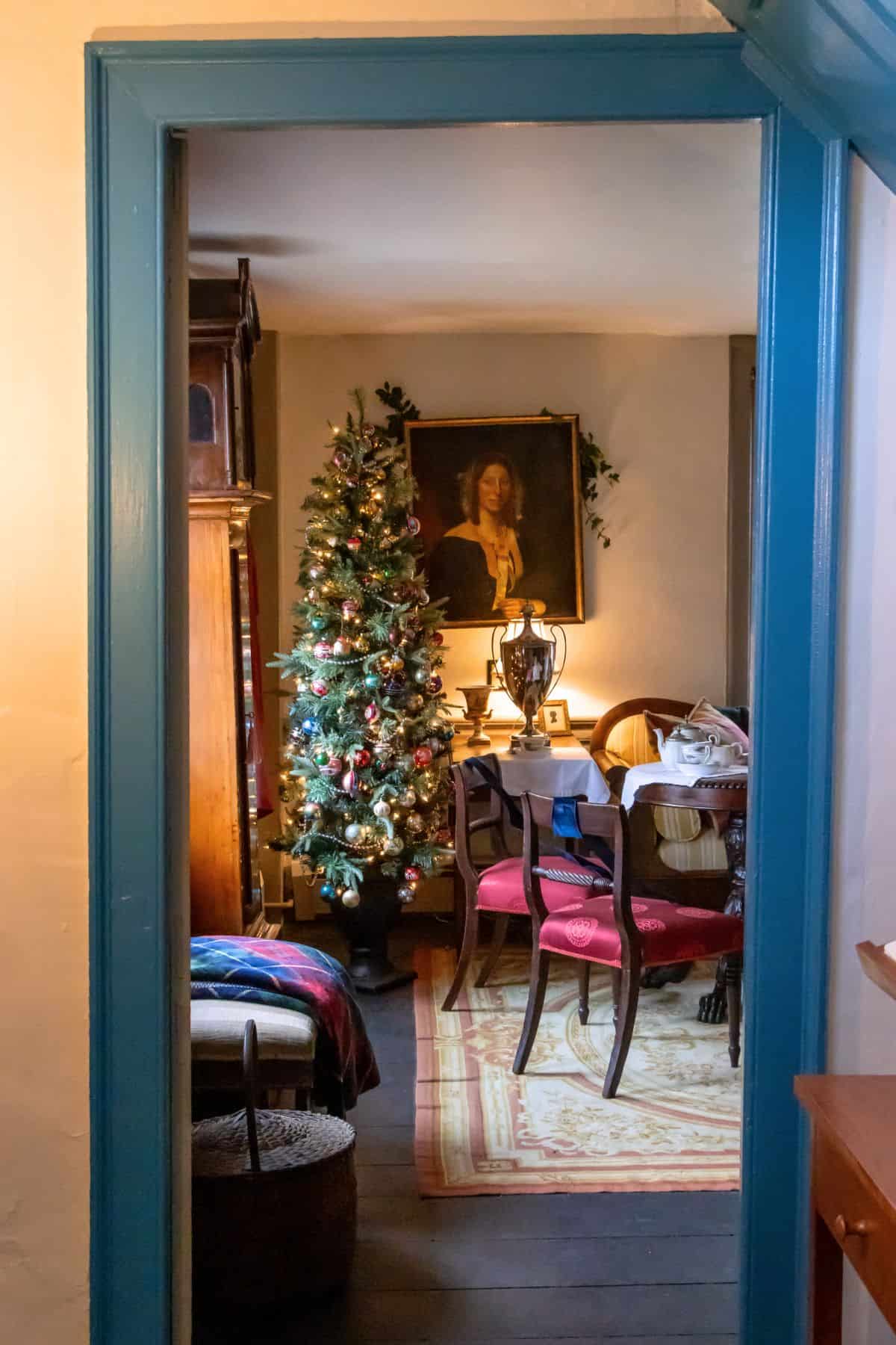 View through a doorway into a warmly lit room with a decorated Christmas tree, a dining table with chairs, a large portrait on the wall, and various household items.