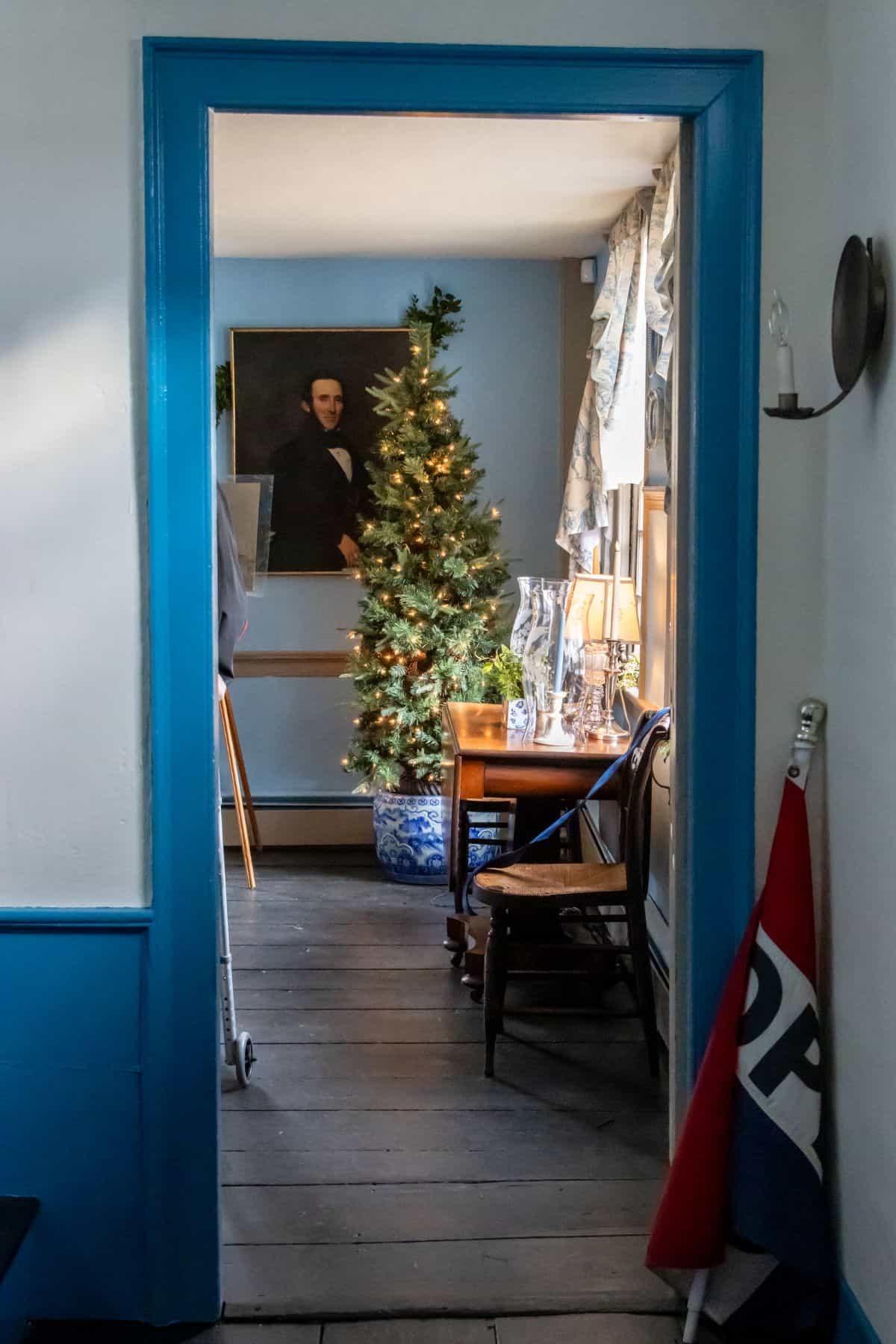 A Christmas tree and a table with a lamp are visible through a doorway with blue trim; a portrait hangs on the wall and a flag is propped against the wall on the right.