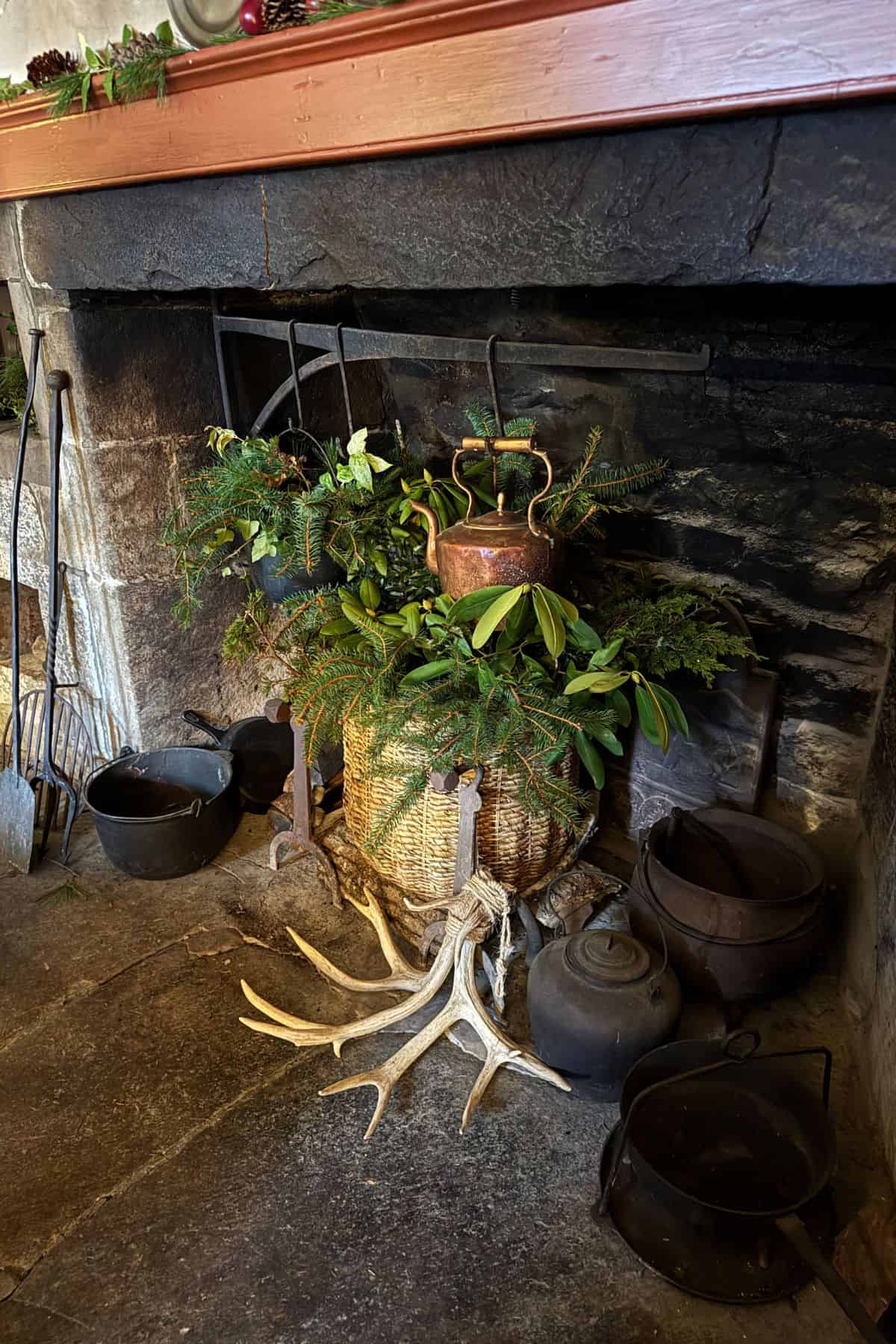 A wicker basket with greenery and a copper kettle sits in a stone fireplace, surrounded by antlers and several cast iron pots.