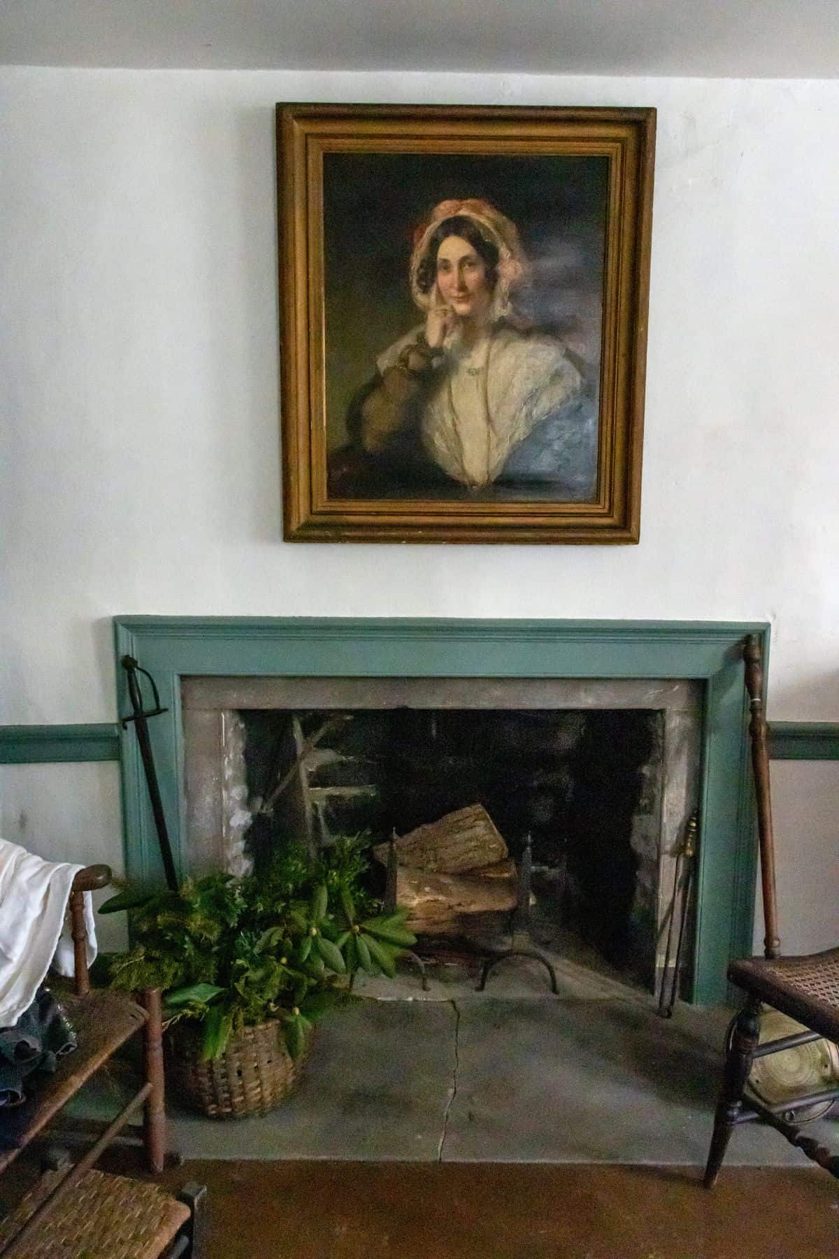 A framed portrait of a woman hangs above a fireplace with logs, a basket of greenery, and wooden chairs on either side.