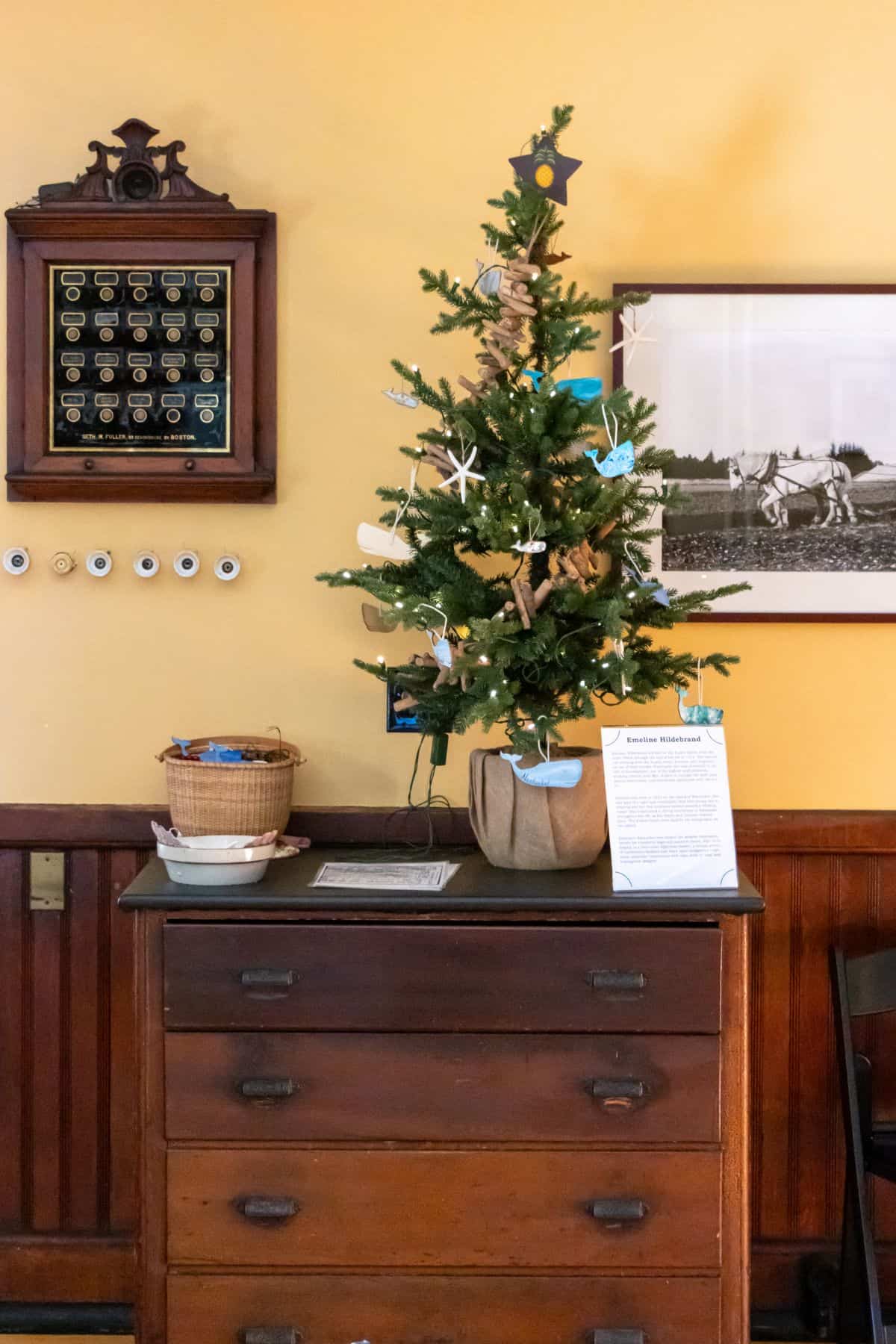 A small decorated Christmas tree sits on top of a wooden dresser, next to a basket and a framed paper, with a photograph and wall plaque hanging above.