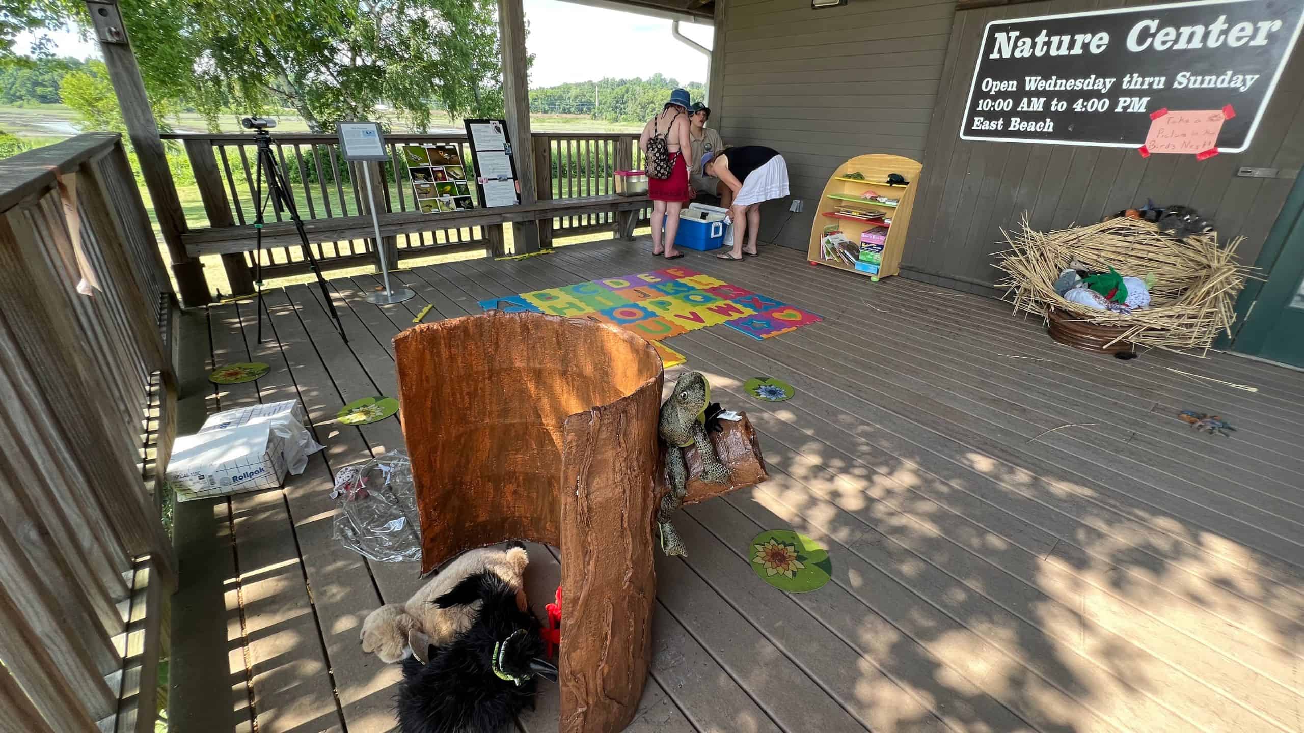 A wooden deck at a nature center with animal displays, a log prop, books, a large mat with letters, and two people looking at items on a shelf.