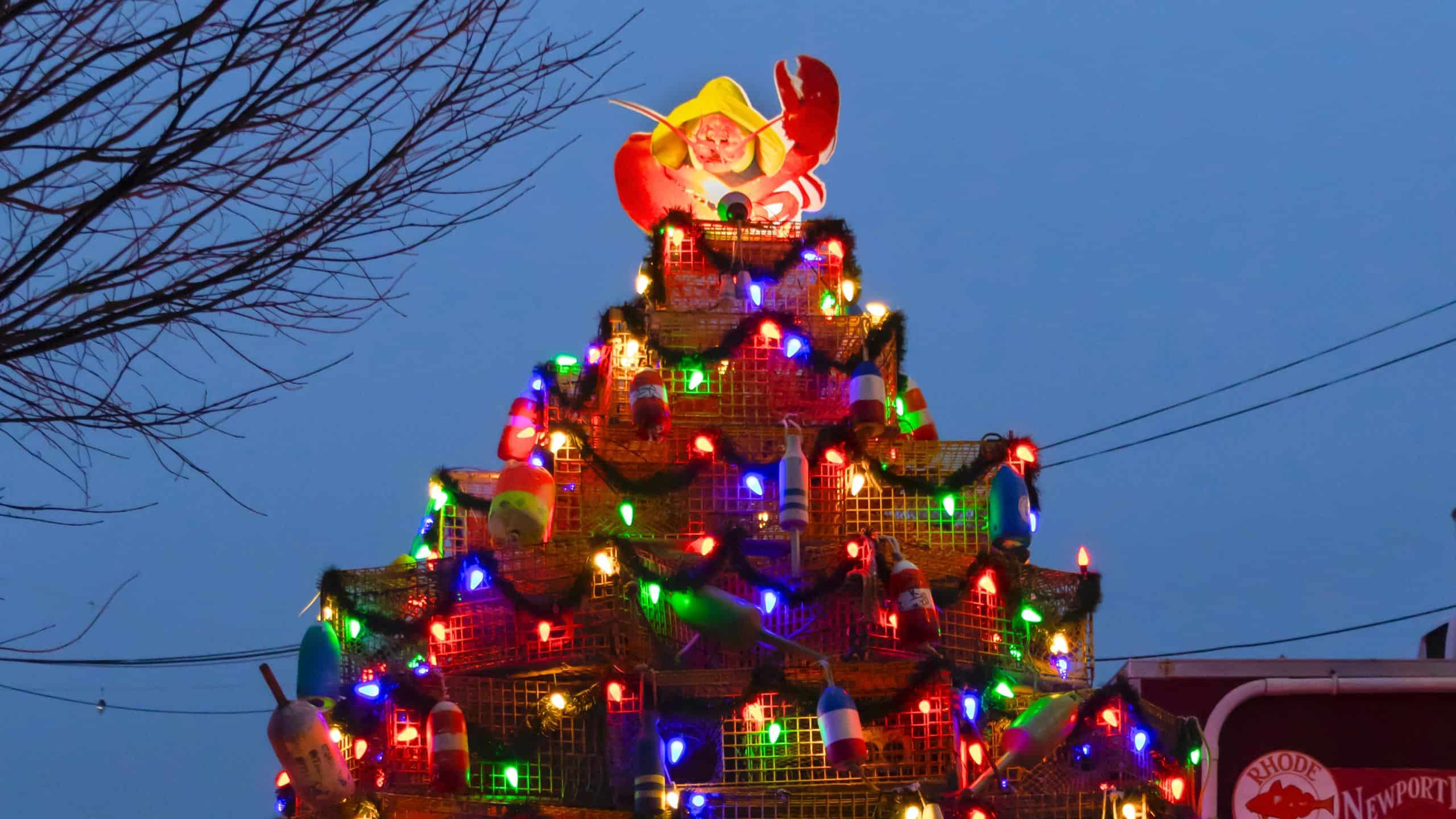 A Christmas tree made of stacked lobster traps decorated with colorful lights and buoys, topped with a Santa Claus holding a lobster, set against an evening sky.