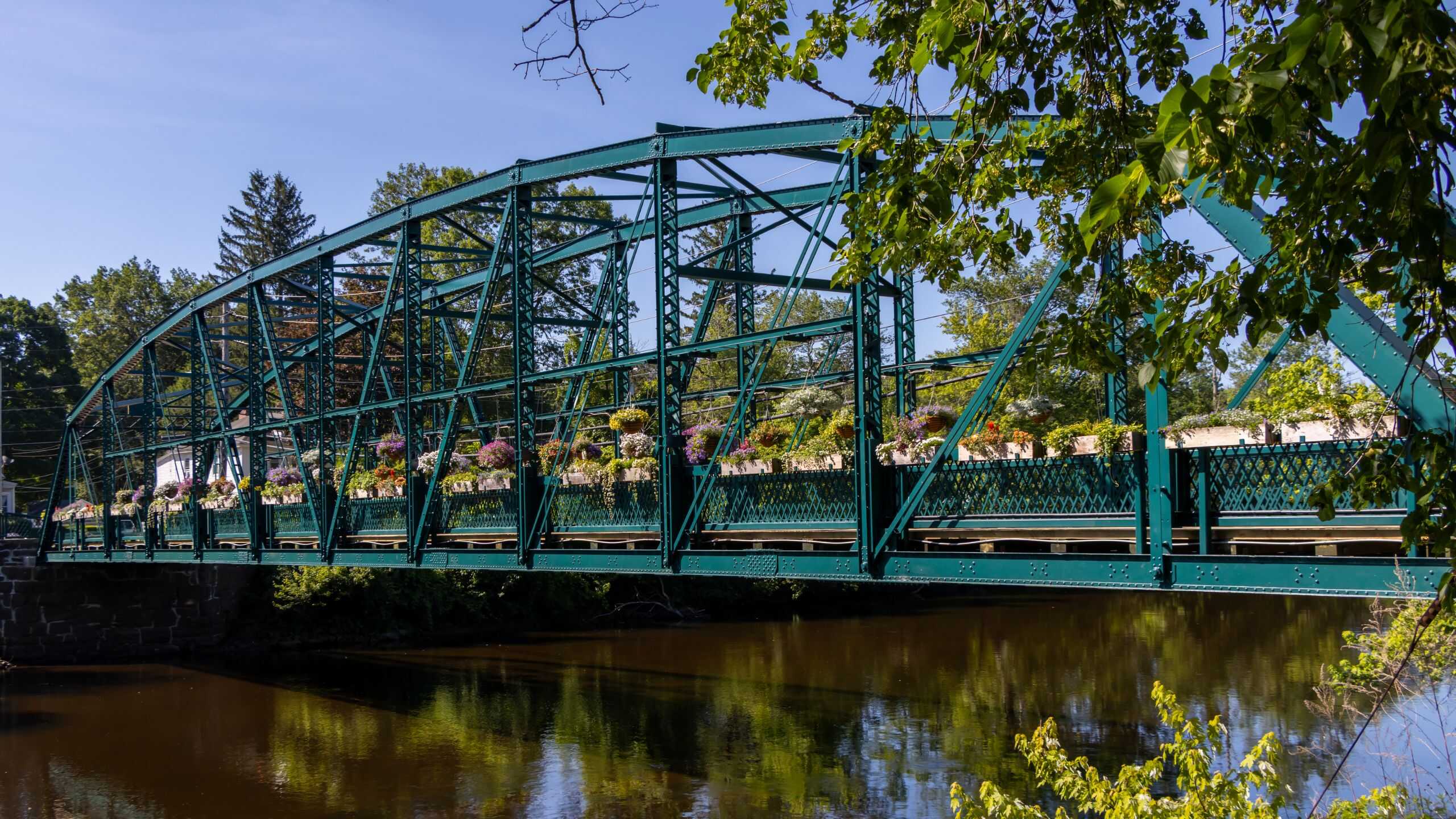 A green metal truss bridge with flower planters crosses over a calm river, surrounded by trees and foliage under a clear blue sky.