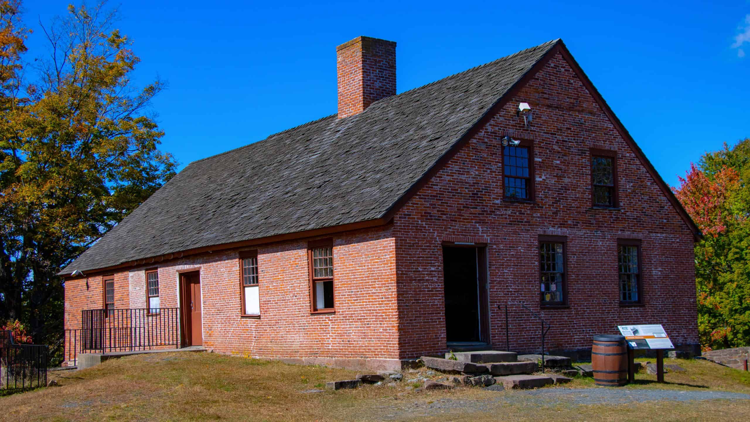 A historic brick building with a steep shingle roof, chimney, and steps leading to the entrance, set against a clear blue sky and autumn trees.