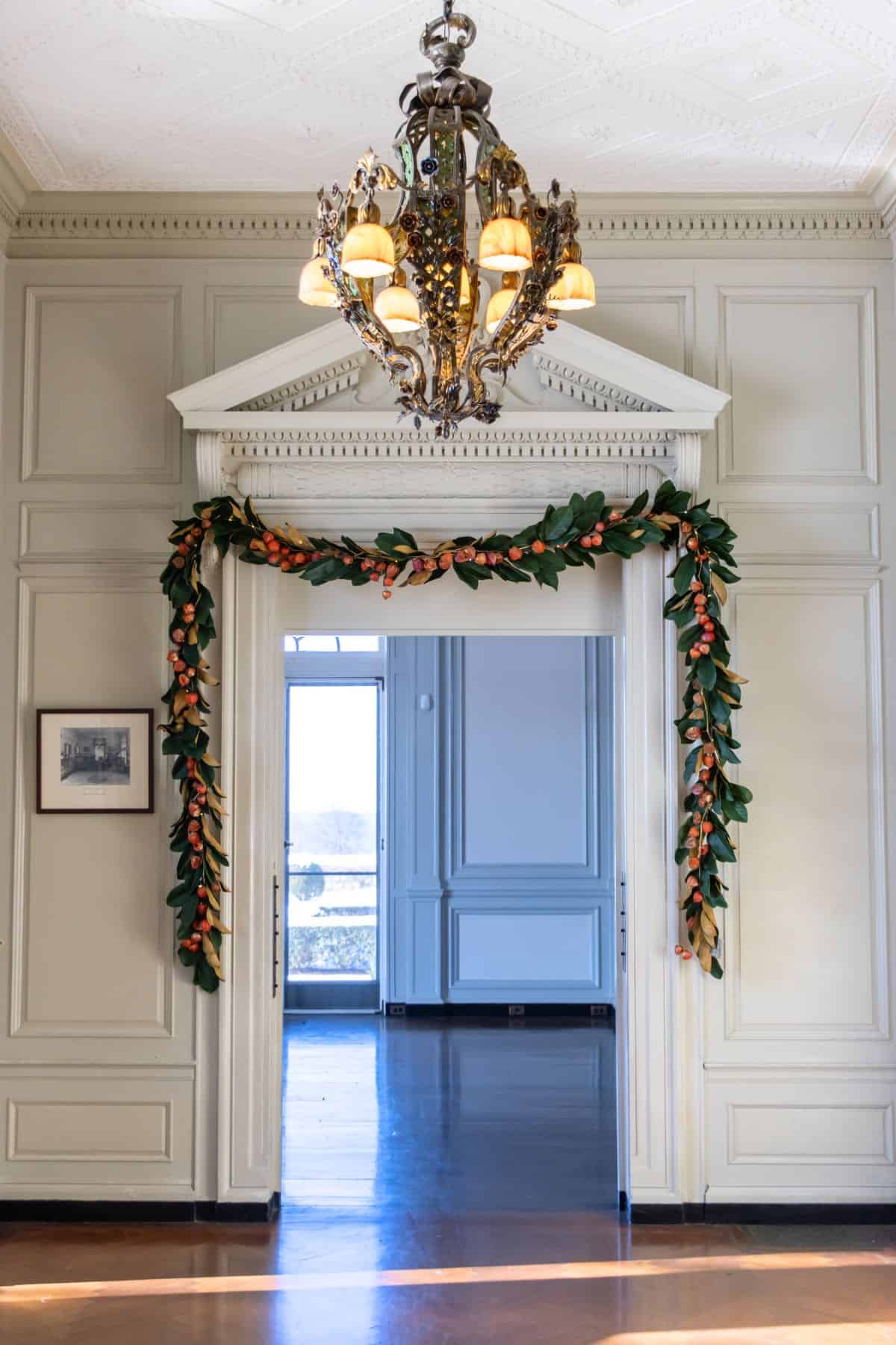 A doorway decorated with a garland of greenery and orange berries, beneath an ornate chandelier, leads into a sunlit room with blue-gray paneled walls.