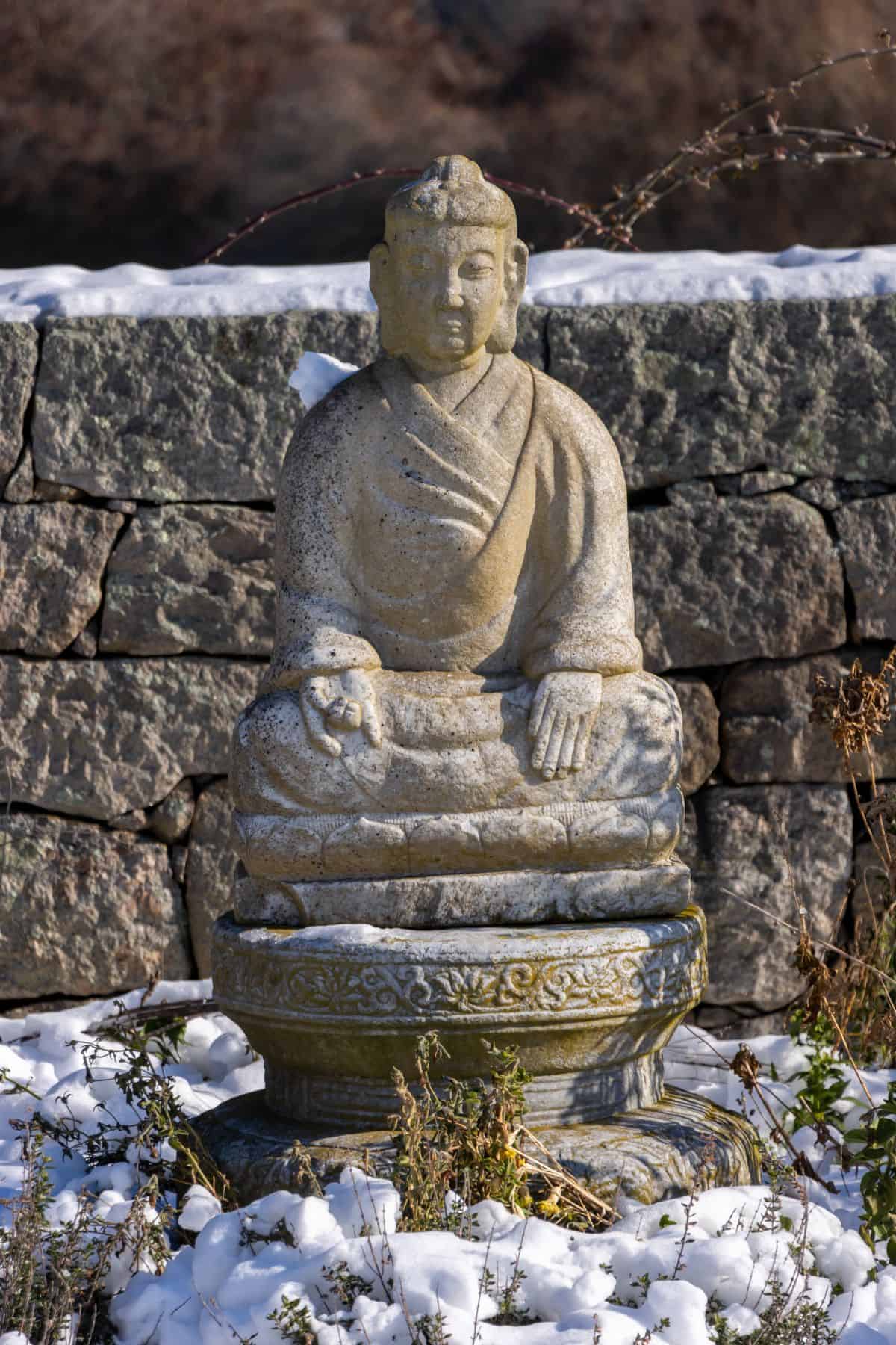 A stone statue of a seated Buddha on a pedestal, surrounded by snow-covered ground and plants, with a stone wall in the background.