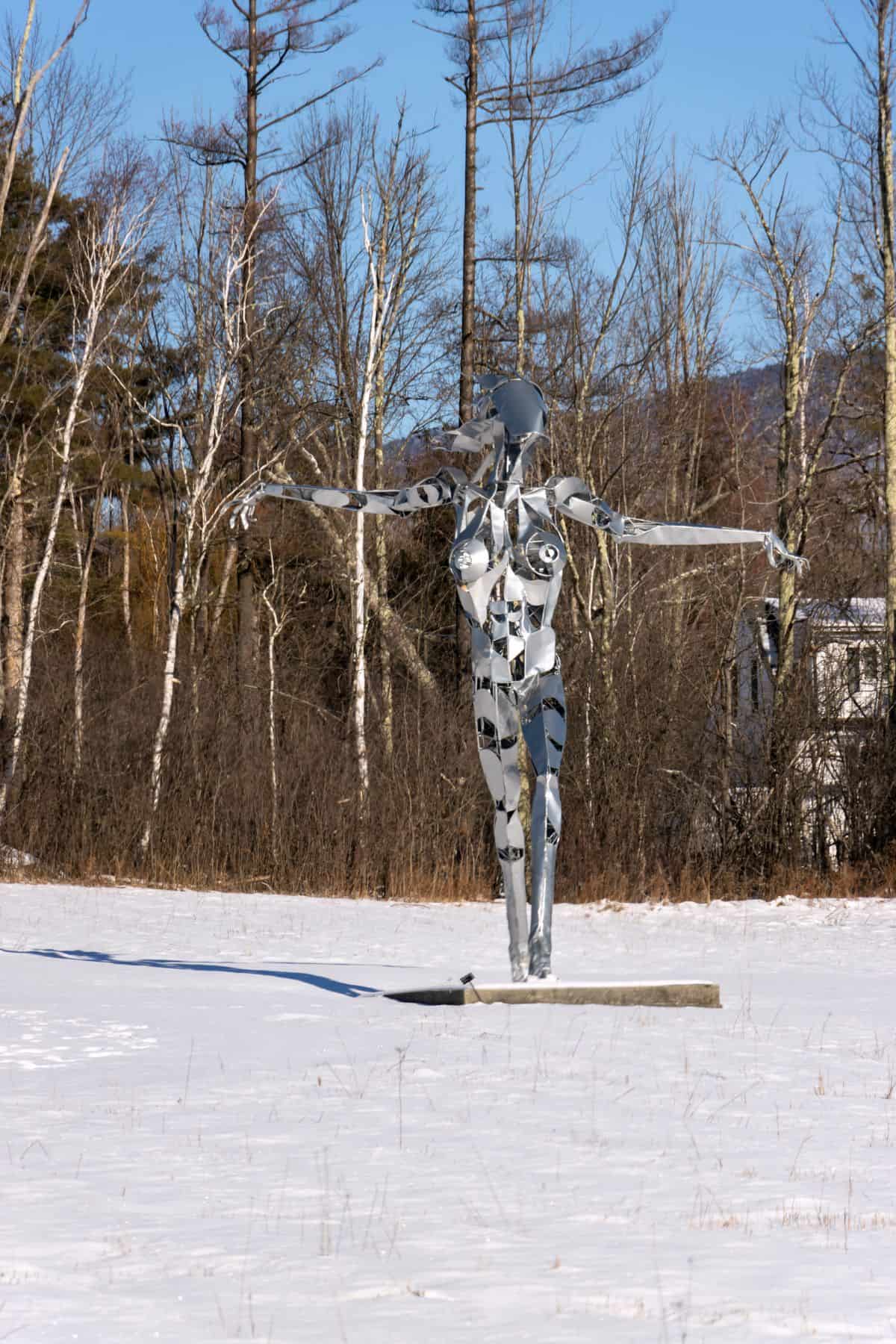 A metallic humanoid sculpture with outstretched arms stands on a snowy field, with bare trees and a clear blue sky in the background.