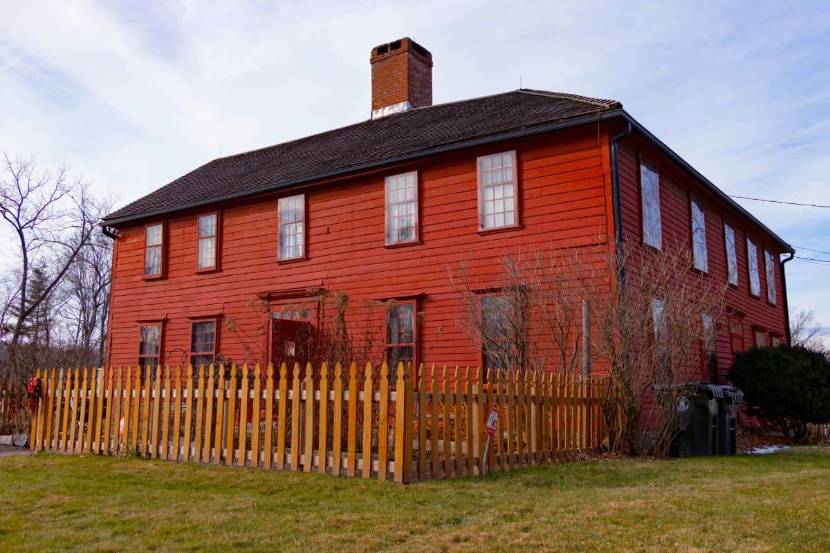 A large, two-story red wooden house with a fenced yard, multiple windows, and a central brick chimney under a blue sky.